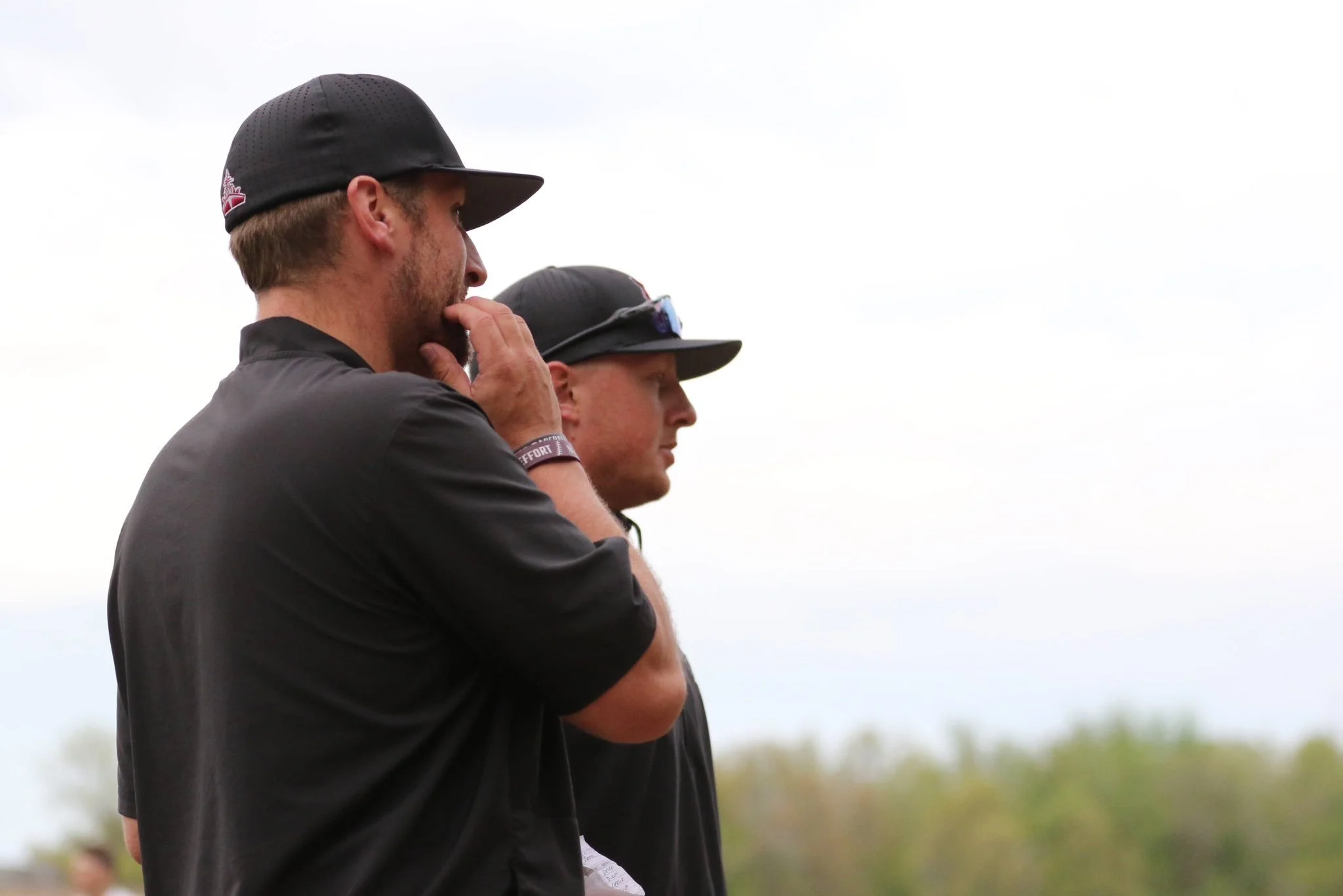 Two men are standing outdoors, wearing dark clothing and hats. One is touching his face thoughtfully, while the other is in the background with a cap and sunglasses, watching, under a cloudy sky.