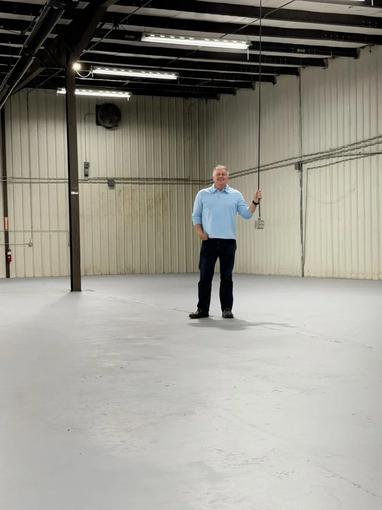 A man standing in an empty warehouse or industrial space, holding a long black pole with a string attached, against beige walls with visible wiring and pipes.