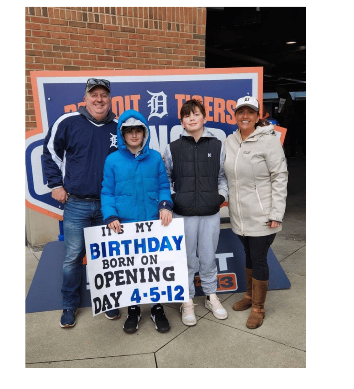 Co-founder Bryan Danek's family at a Tigers game, his son holding a birthday sign. The sign reads 'It's my birthday born on opening day 4-5-12' and they are dressed in casual clothing.