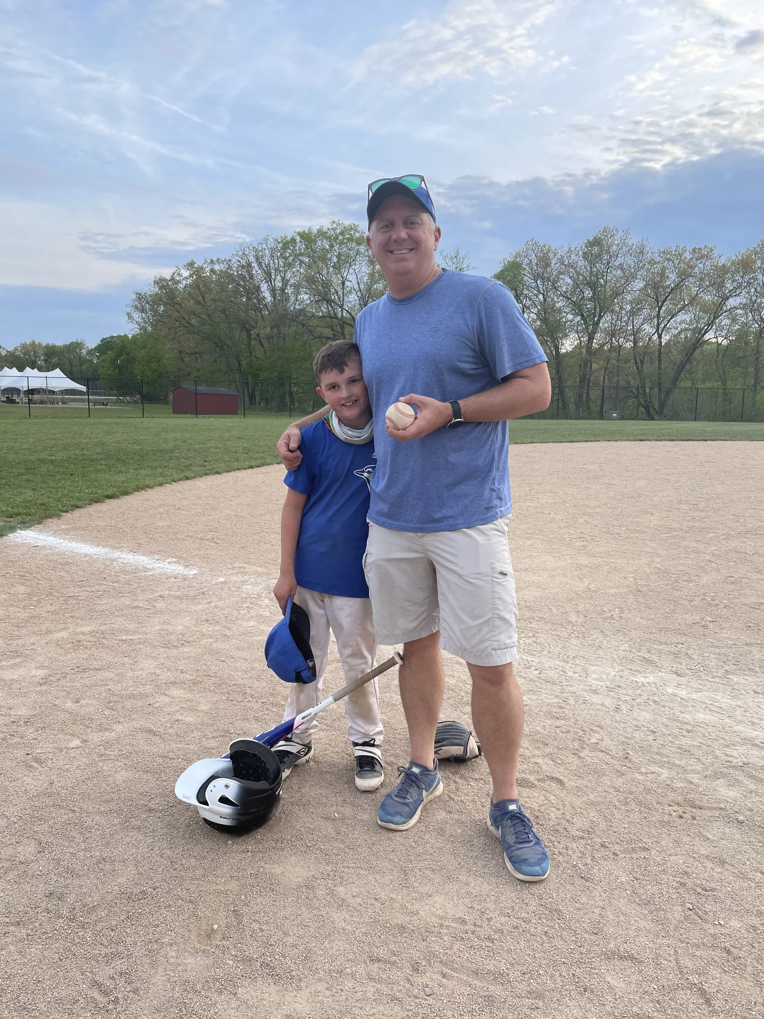 Bryan Danek Co-founder standing with his son on a baseball field, smiling. The man is holding a baseball, and there is a helmet and a scooter on the ground nearby. In the background, trees and a fence are visible under a partly cloudy sky.
