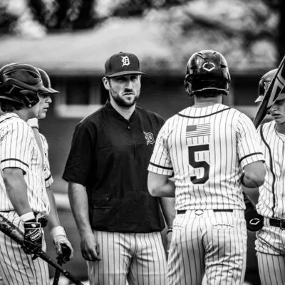 Baseball coach talking to players during a game, all in striped uniforms and helmets, on a baseball field.