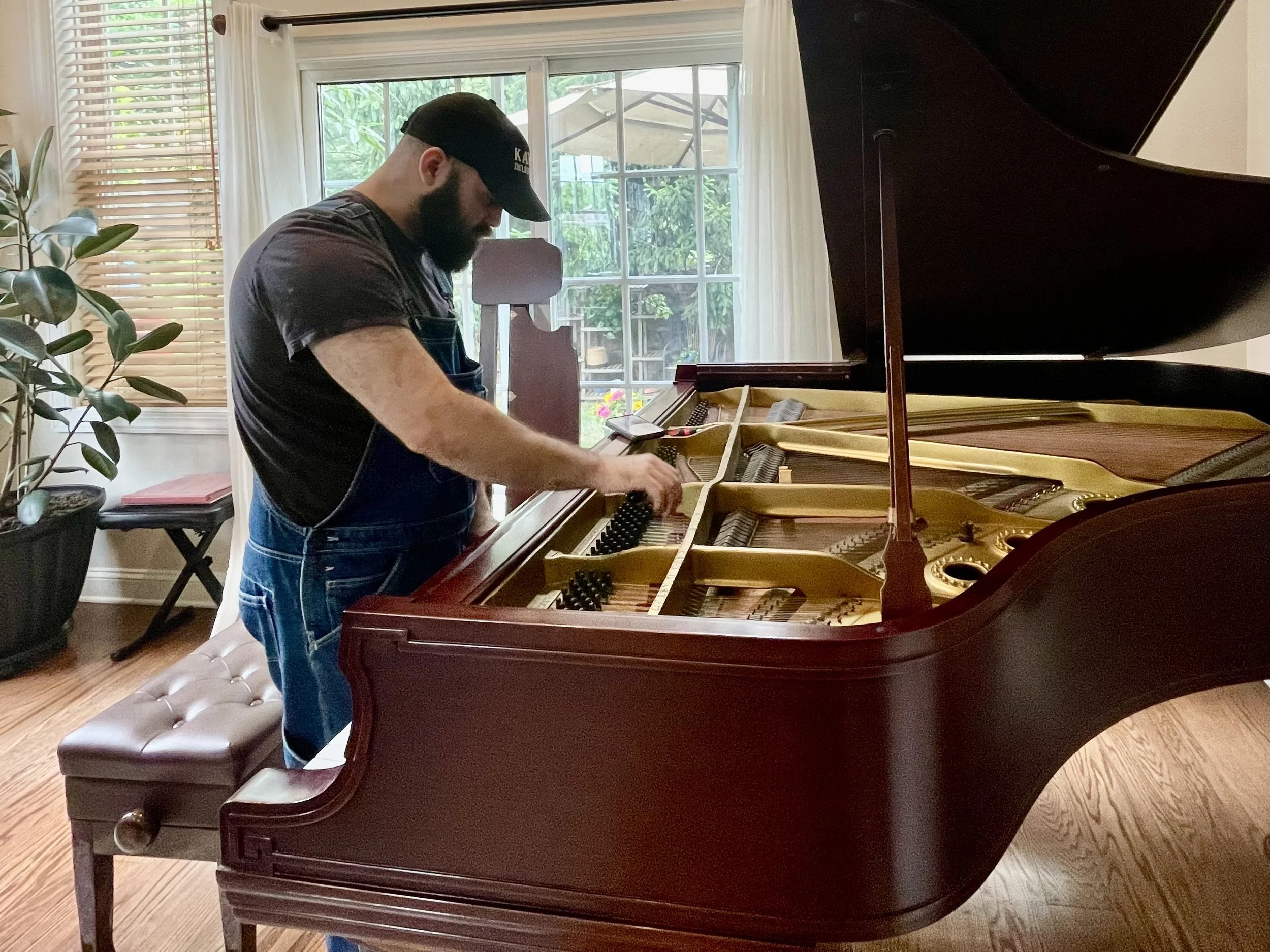 A man with a beard wearing a black cap and apron playing a grand piano in a bright room with large windows and curtains.