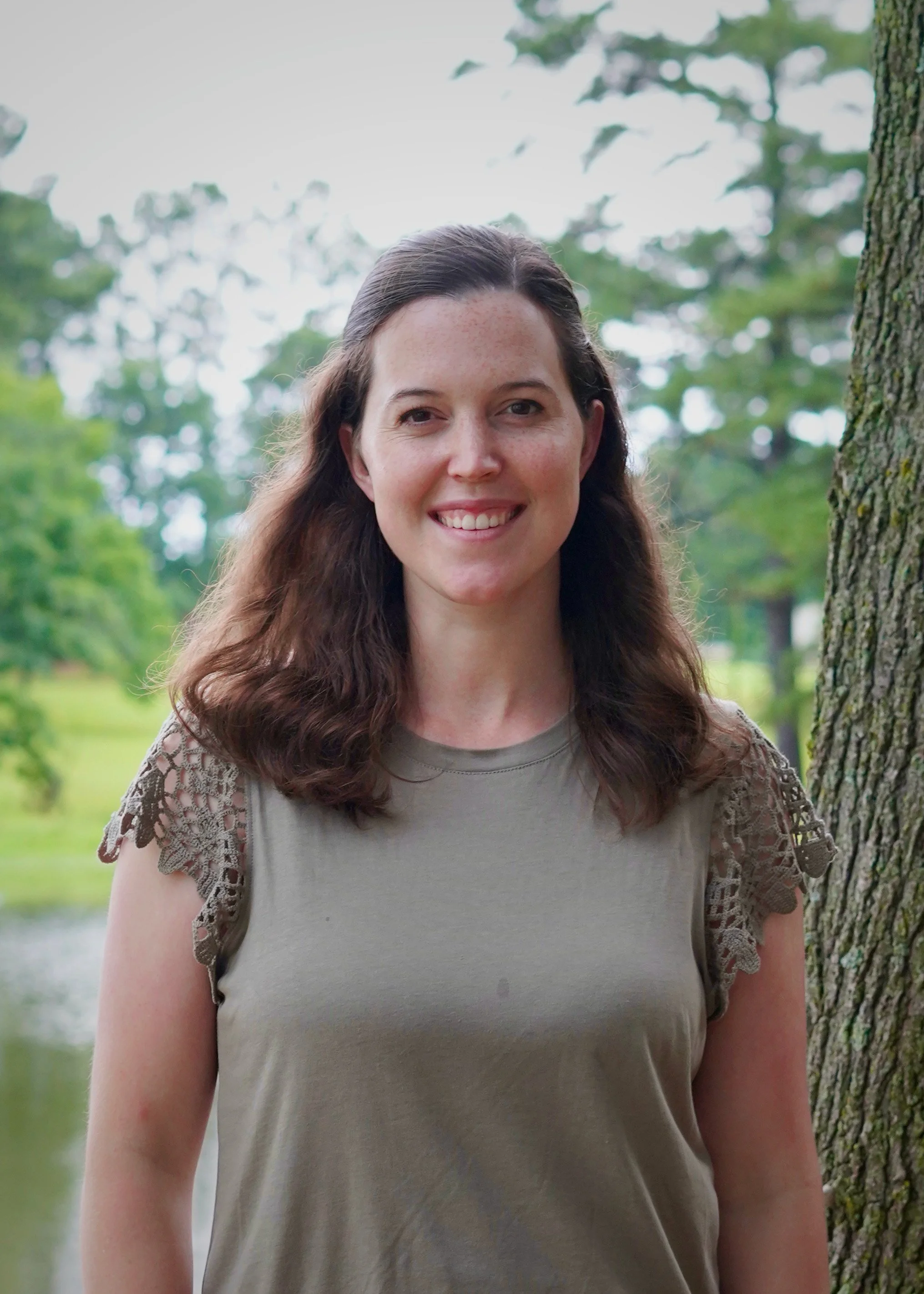 A woman with long brown hair, wearing a beige top with lace sleeves, standing outdoors near a tree with a blurred background of green trees and a body of water.