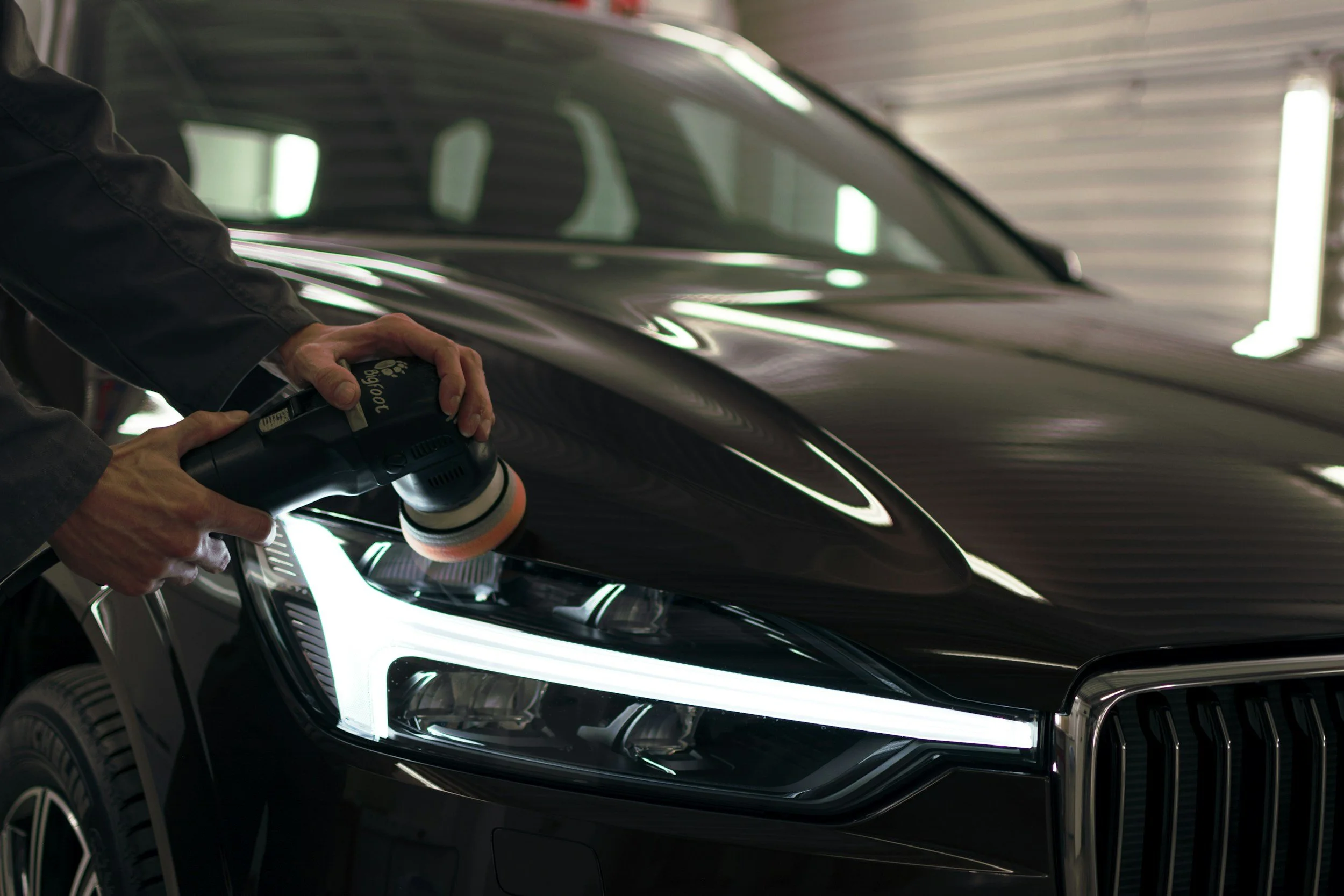 Person polishing the front headlight of a black car in a garage using a polishing machine.