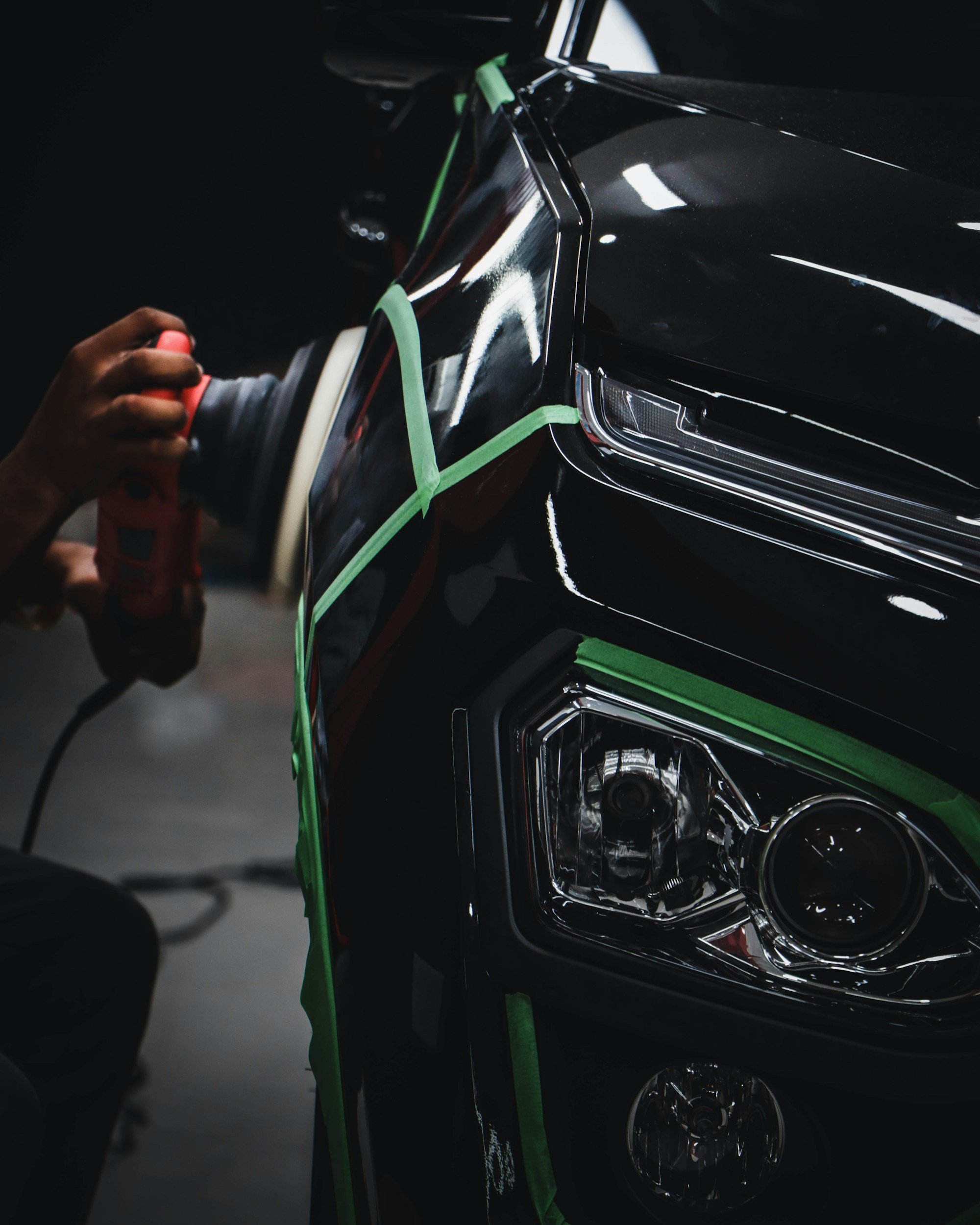 Close-up of a car being polished, with green painter's tape around the headlight and fender, and a hand holding a polishing tool.