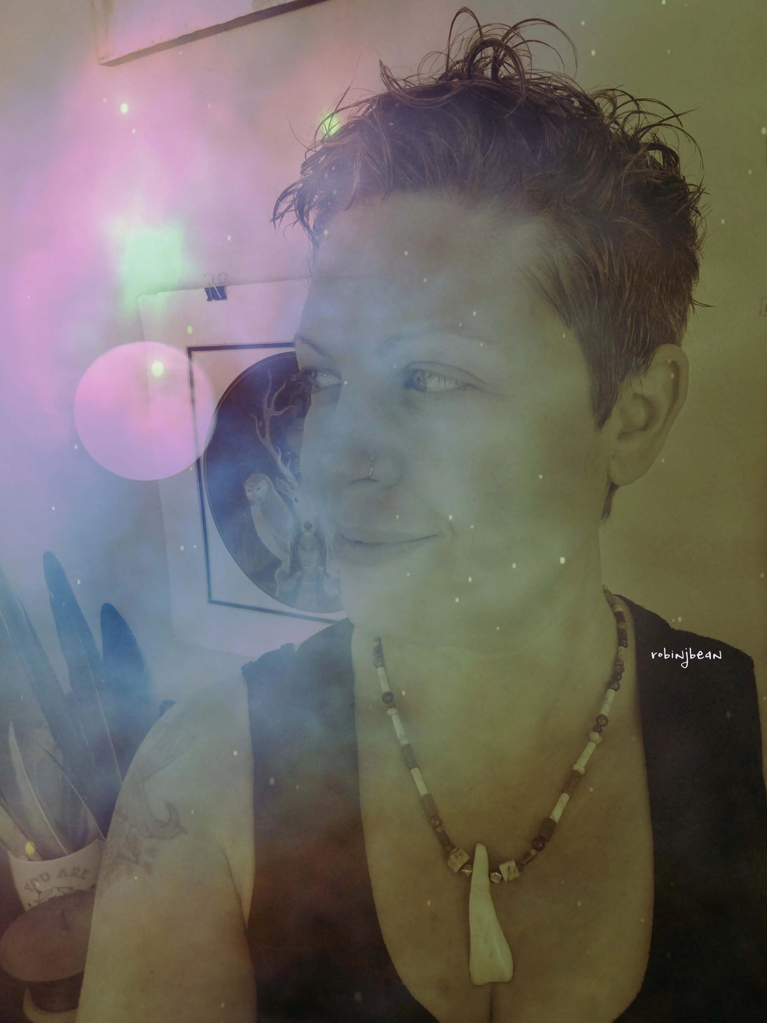 A person with short, curly hair wearing a beaded necklace and a septum piercing, leaning against a wall with their eyes looking to the side. There are some art or posters in the background.