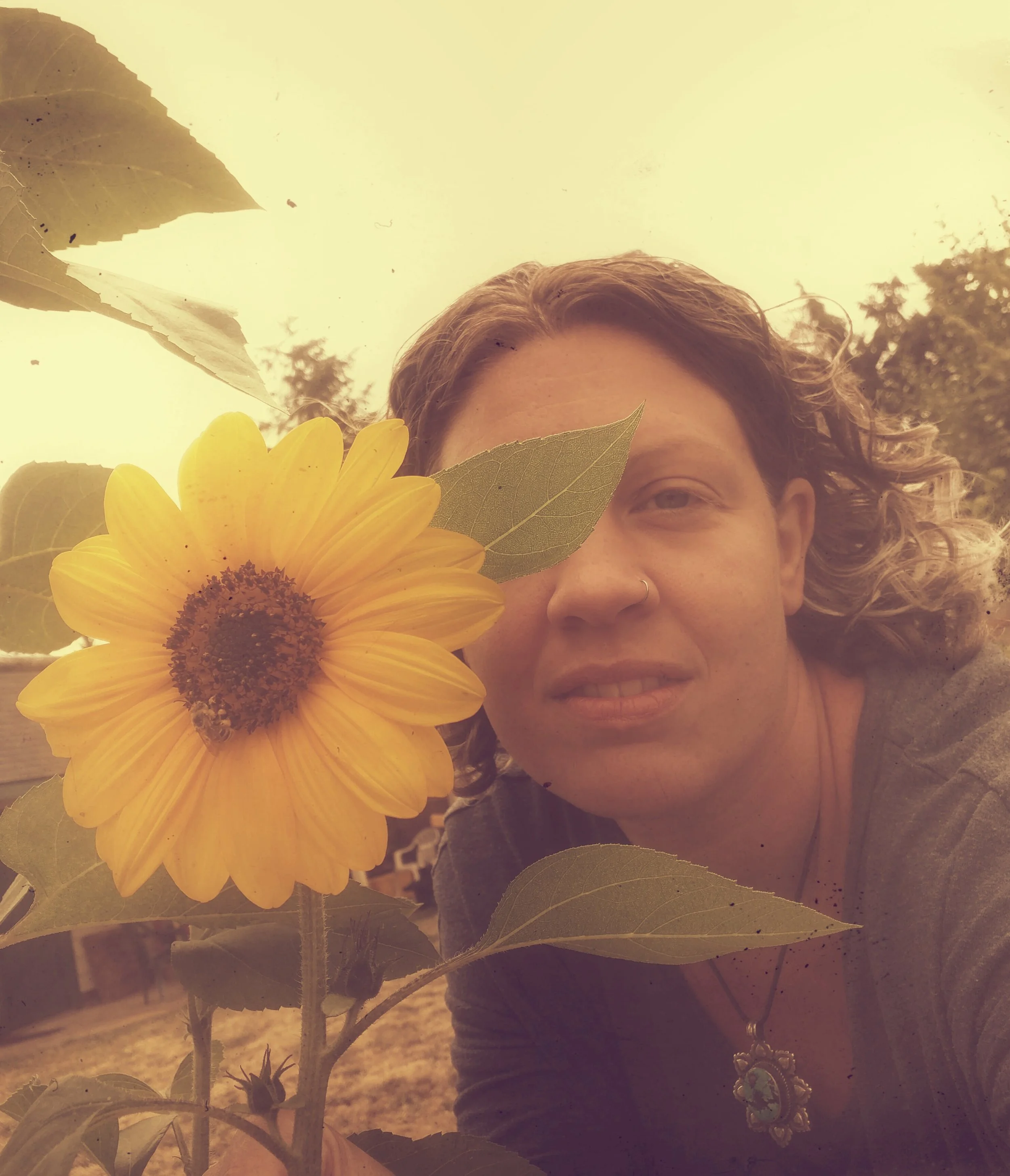 A woman with curly hair holding a large yellow sunflower close to her face in an outdoor setting at sunset.
