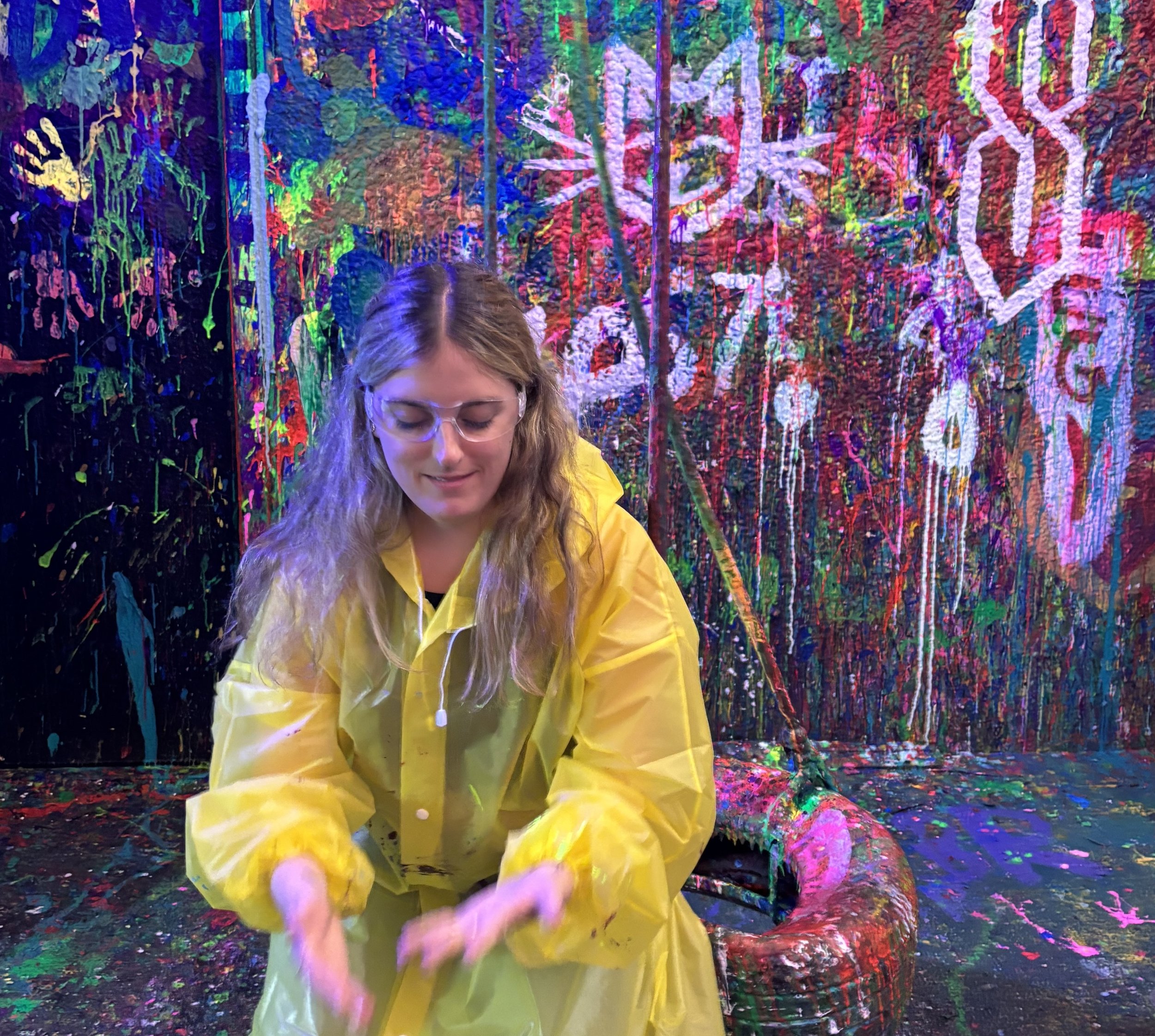 A woman with long blonde hair and glasses wearing a yellow rain jacket, standing in front of a colorful abstract artwork with splashes of bright colors and the word "LOVE" visible in the background.