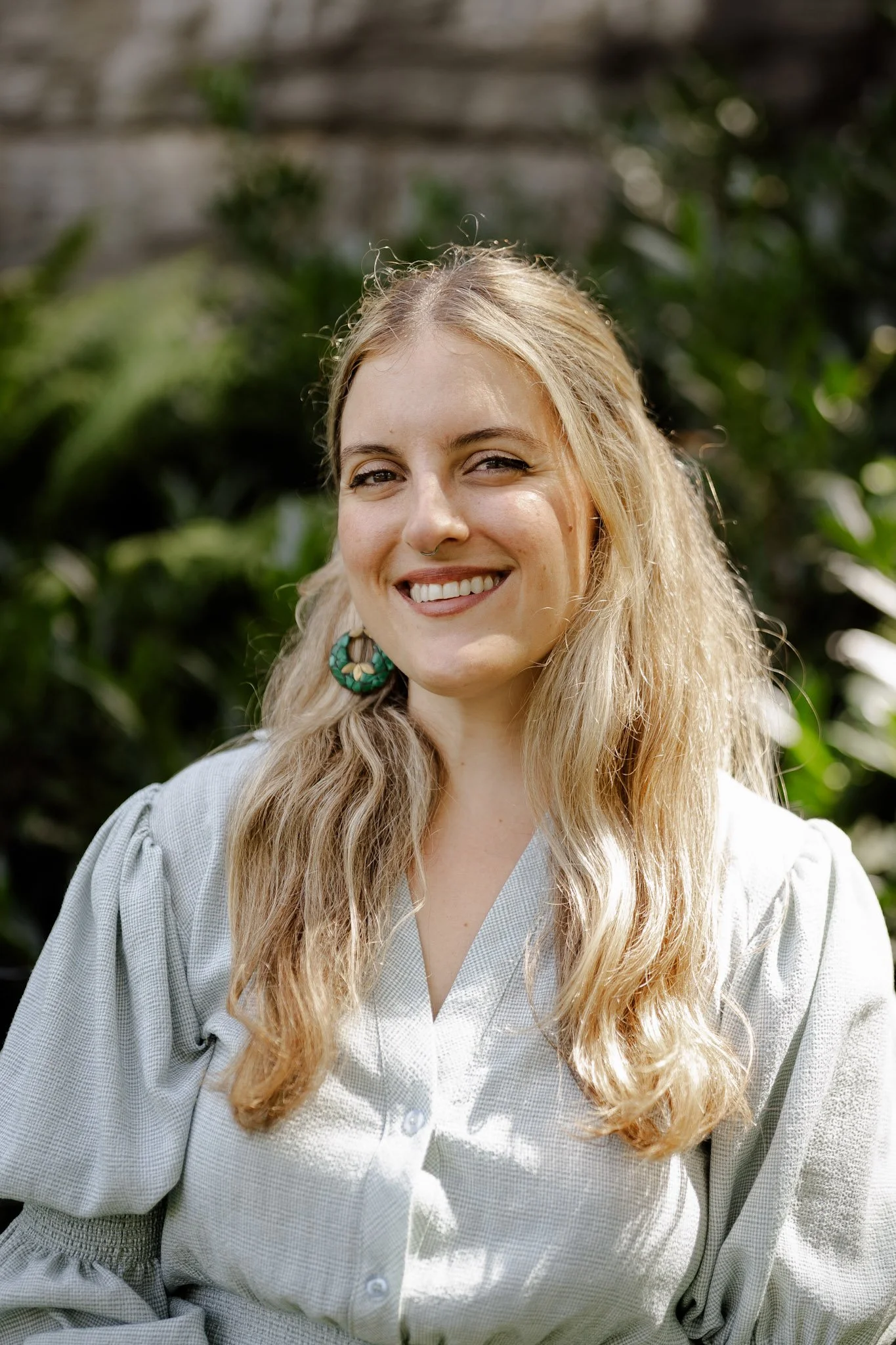 A smiling woman with long wavy blonde hair, wearing a light-colored, loose-fitting blouse with puffed sleeves, standing outdoors with a blurred background of green foliage.