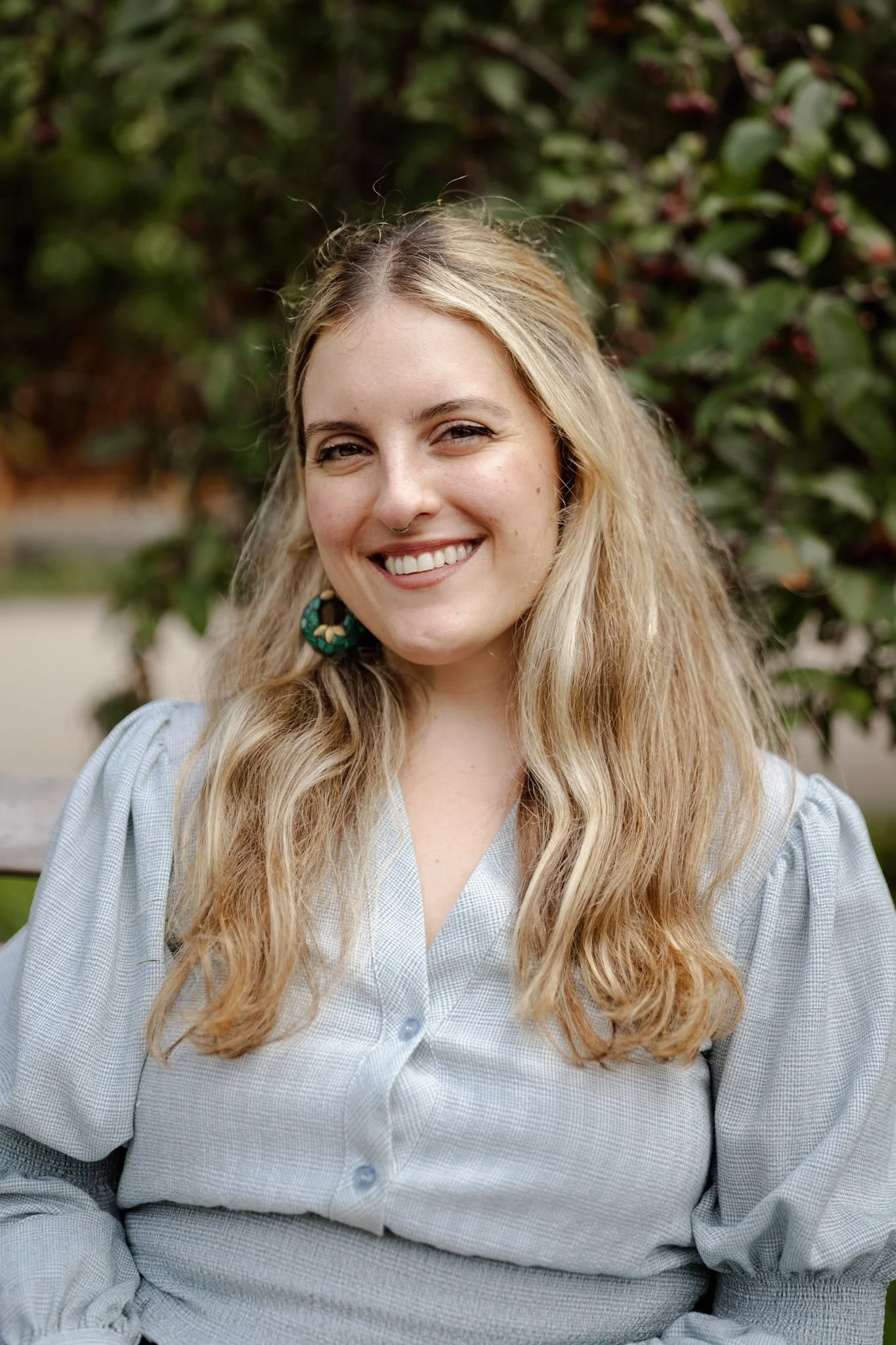 A young woman with long, wavy blonde hair smiling outdoors against a background of green foliage.