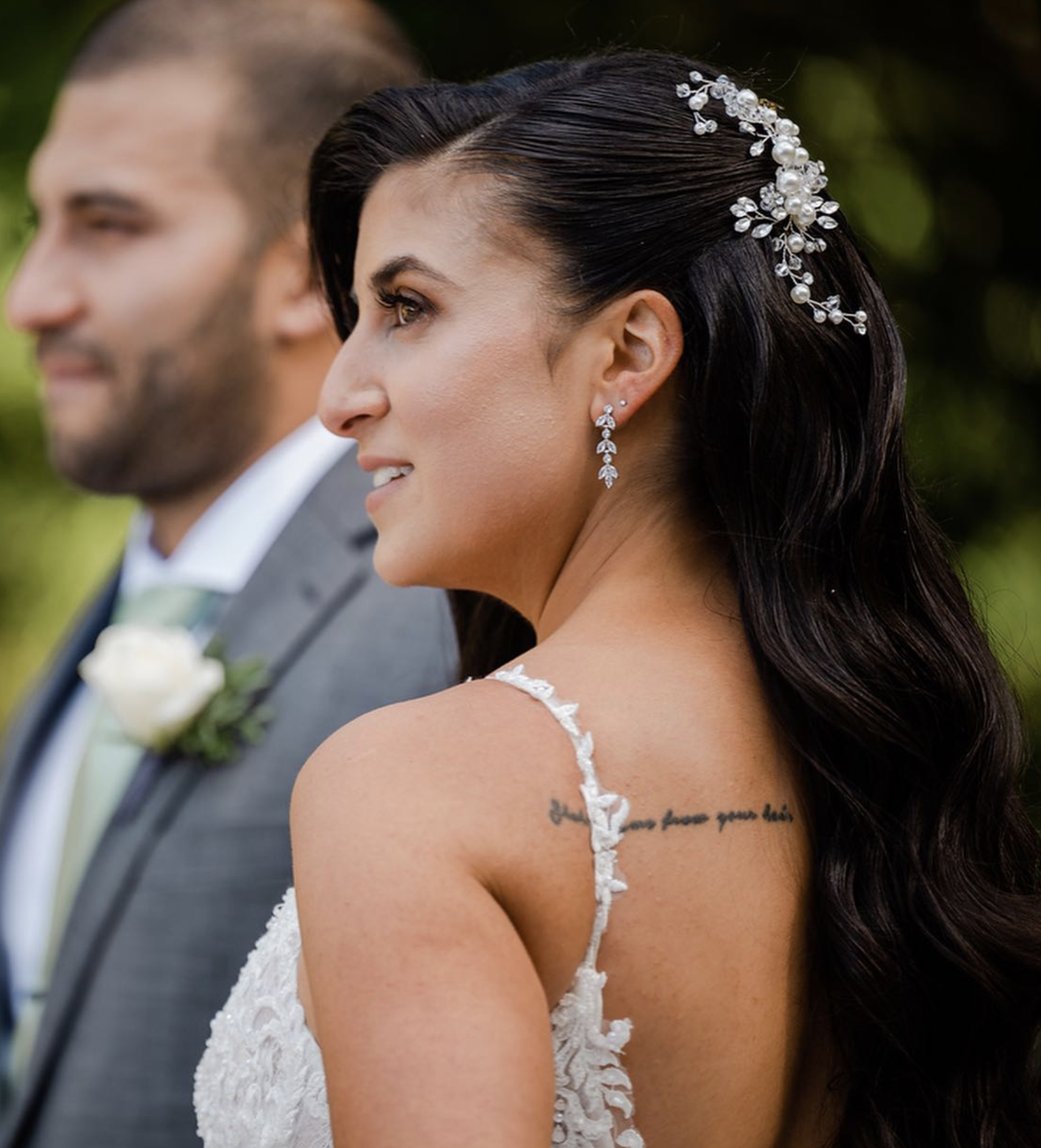 Close-up of a woman with long, dark hair wearing a pearl and rhinestone hair accessory, earrings, and a lace dress, standing next to a man in a suit. The woman has a tattoo on her shoulder and is smiling softly, with a blurred man in a gray suit with a bow tie and boutonniere in the background.