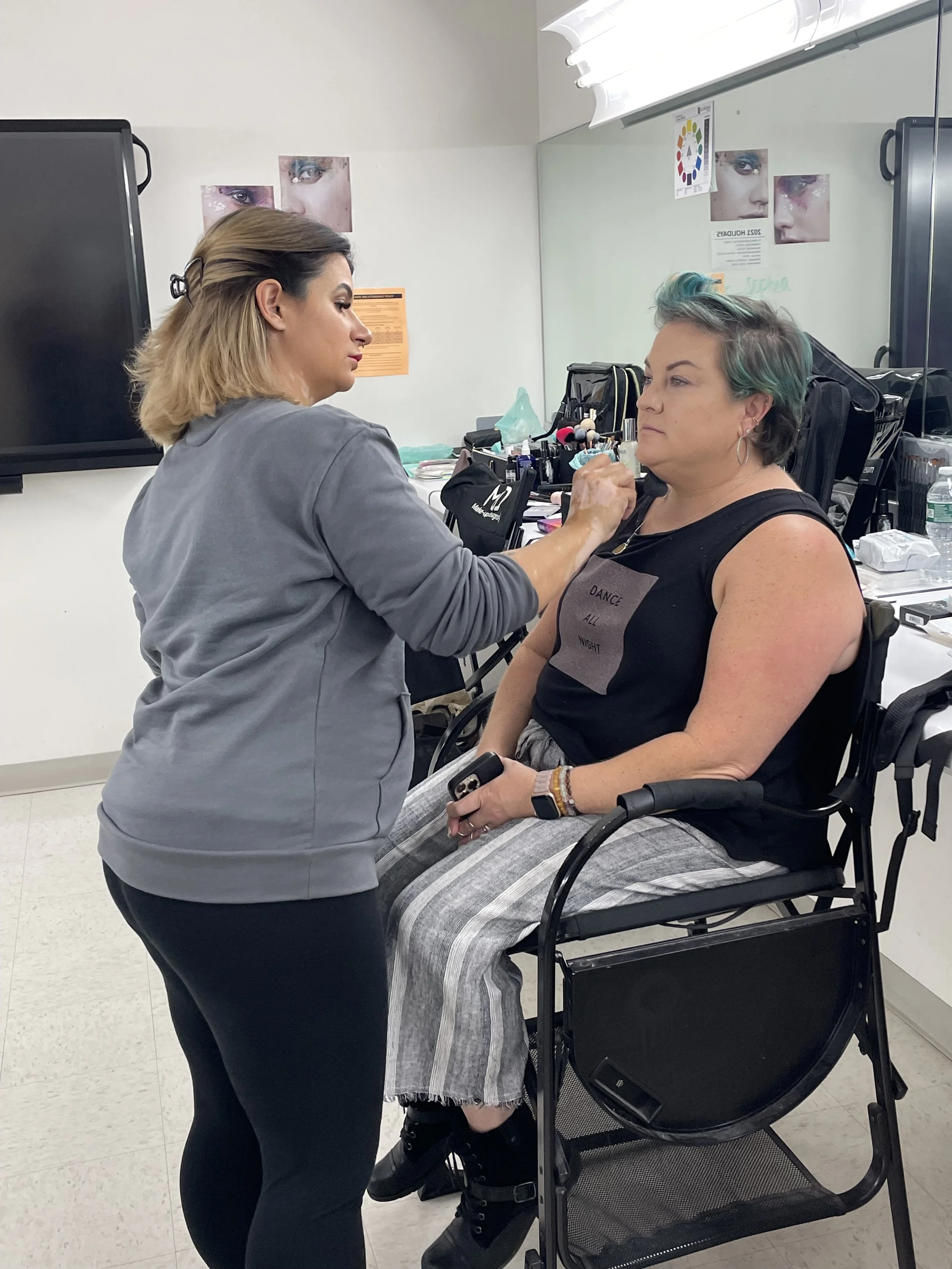 A woman with short, teal and gray hair sitting in a wheelchair while another woman applies makeup to her face in a room with makeup tools and posters on the wall.