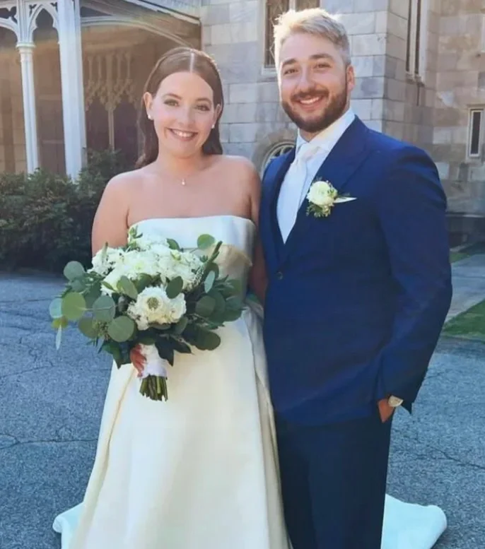A bride and groom standing outdoors, smiling. The bride is holding a bouquet of white flowers and wearing a strapless white wedding gown. The groom wears a dark blue suit with a white shirt and a boutonnière. Behind them is a stone building.