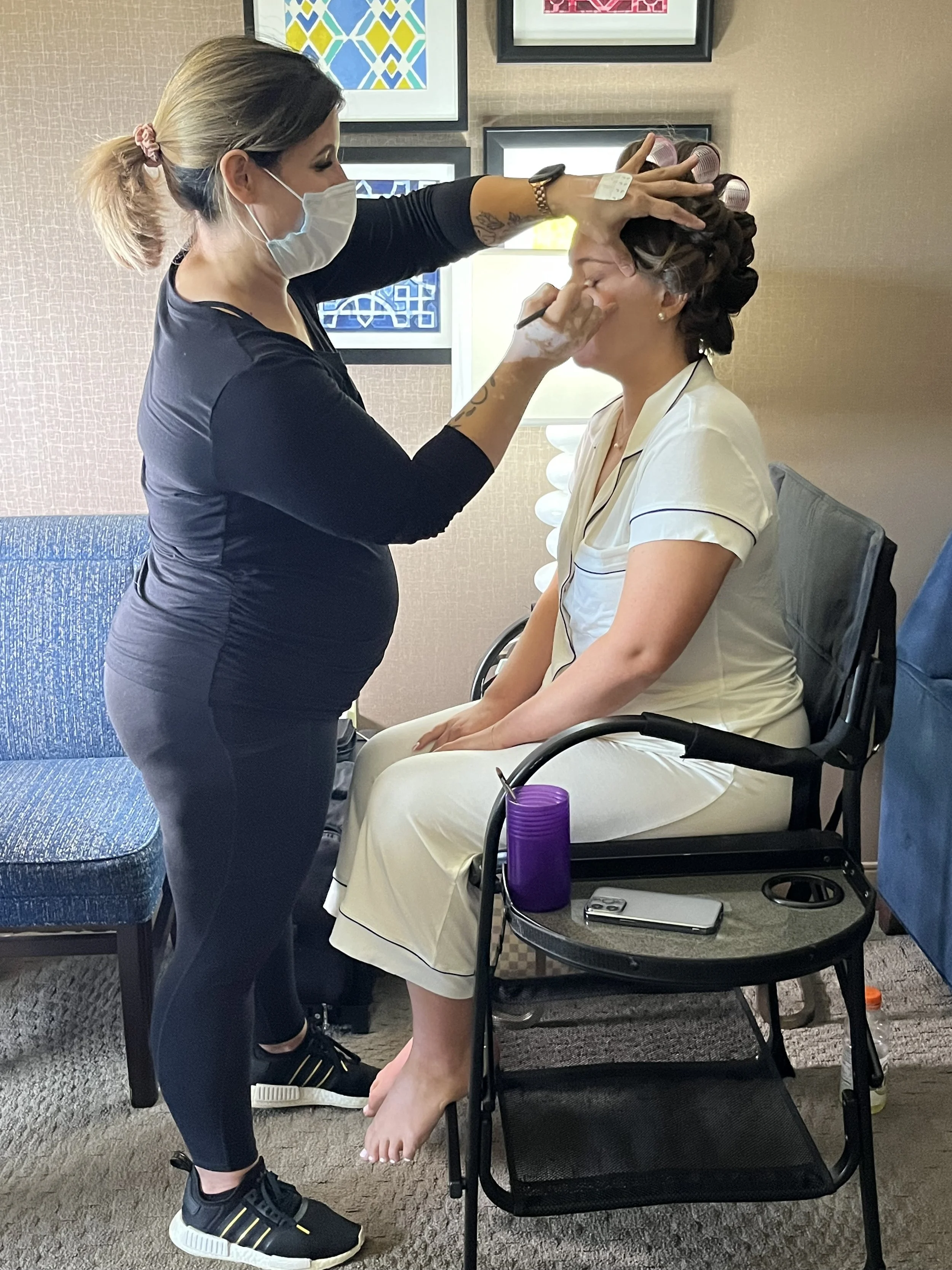 A woman sitting in a chair getting her makeup done by another woman in a black top, wearing a face mask, in a room with framed artwork on the wall. The woman is wearing a white top with black piping and has rollers in her hair. There is a purple water bottle and a cellphone on a small black table beside her.