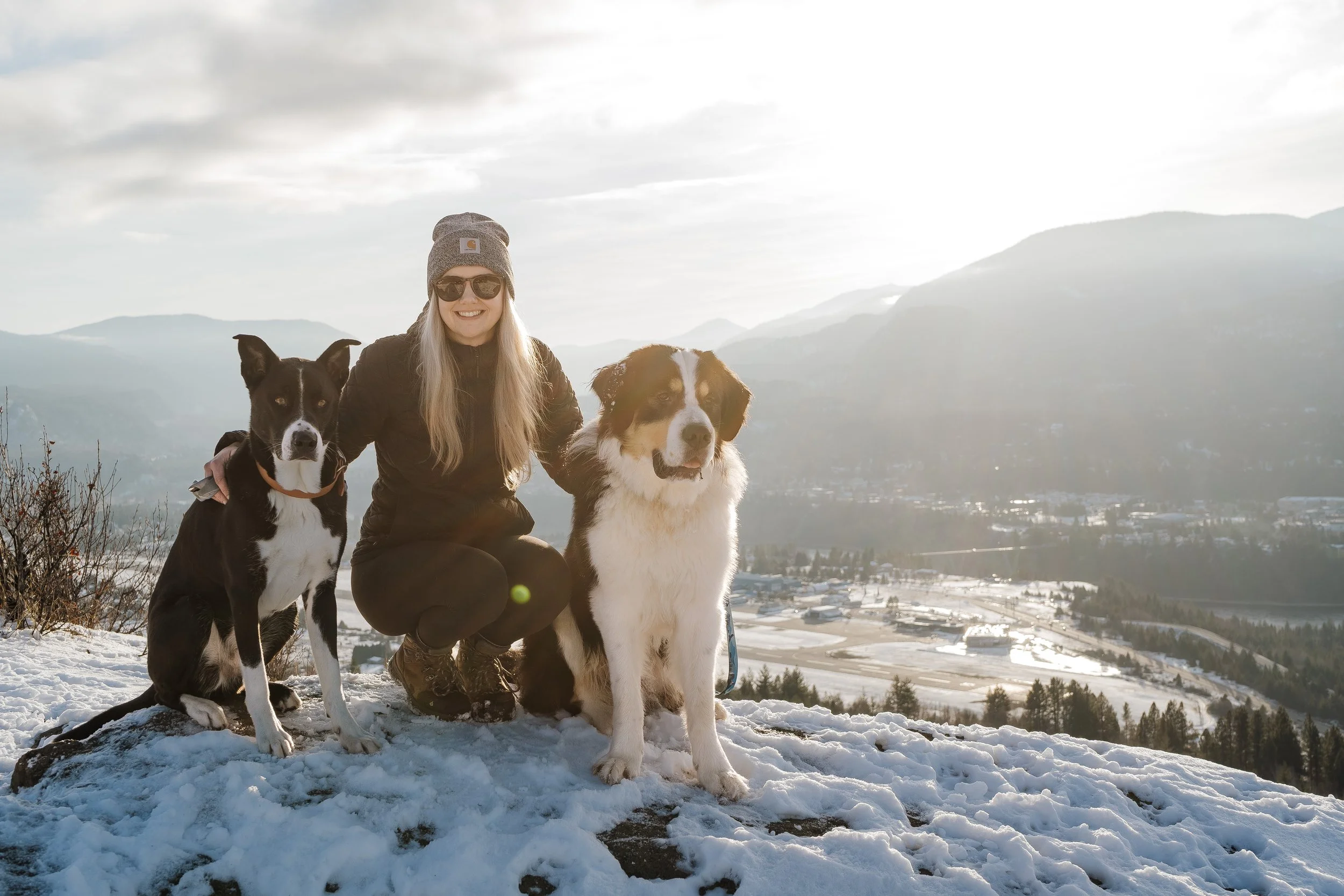 A woman in winter clothing kneeling on snow-covered ground with two dogs, mountains and cloudy sky in the background.
