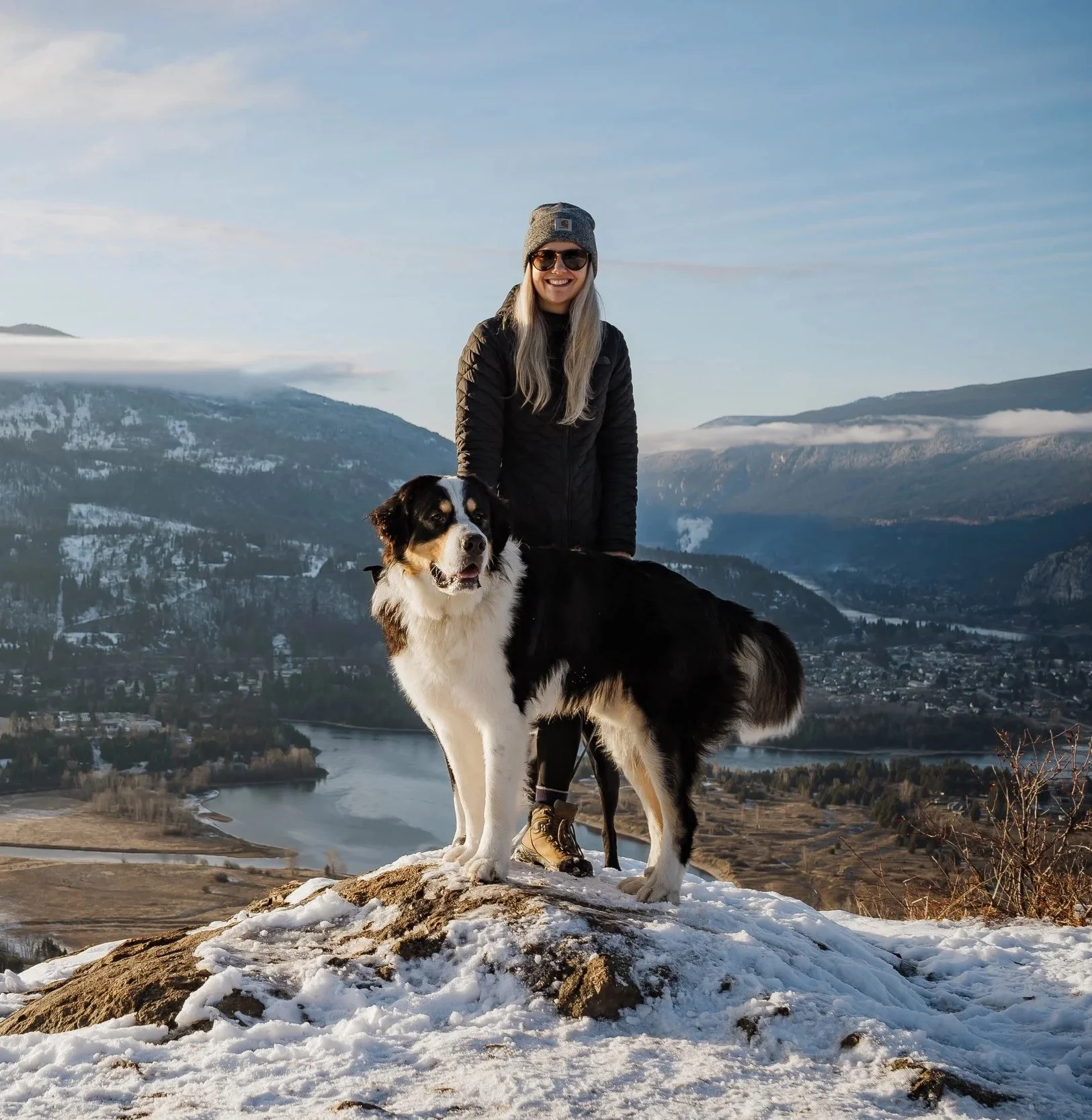 A woman with long blonde hair, sunglasses, and a gray beanie, wearing a black jacket and hiking boots, standing with a black and white dog on a snow-covered hilltop with a river, mountains, and a partly cloudy sky in the background.