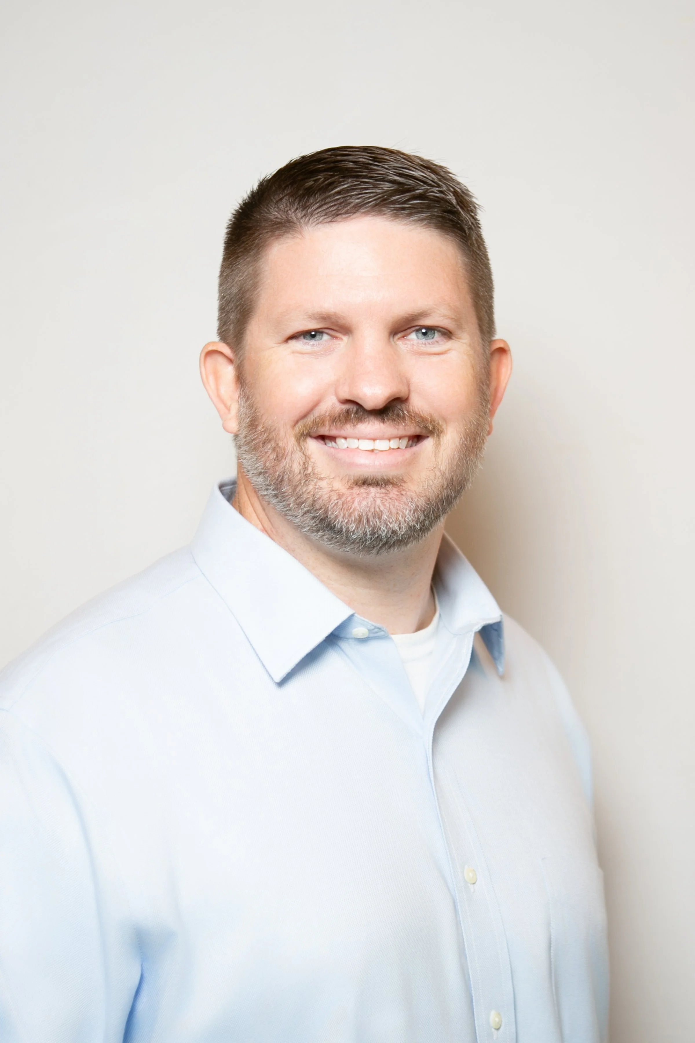 Headshot of a man with short dark hair, light beard, wearing a light blue button-up shirt, smiling, against a plain light background.
