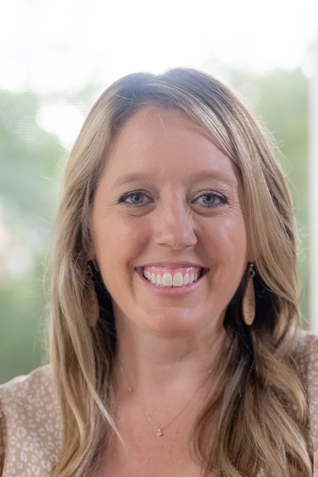 Close-up of a smiling woman with blonde hair, blue eyes, wearing earrings, and a light-colored patterned top, with a blurred outdoor background.