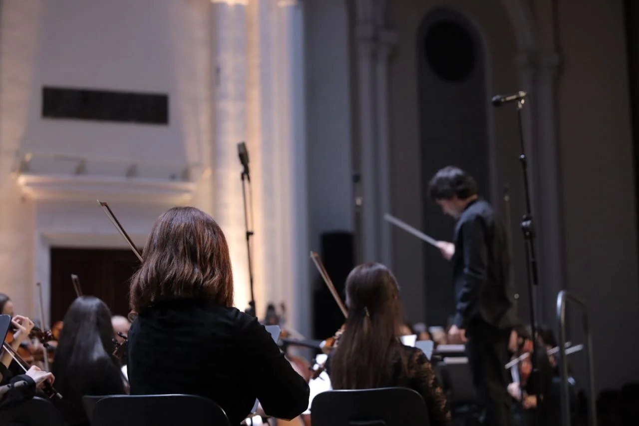 An orchestra conducting a performance in a concert hall, with musicians playing string instruments, and a conductor leading them with a baton.