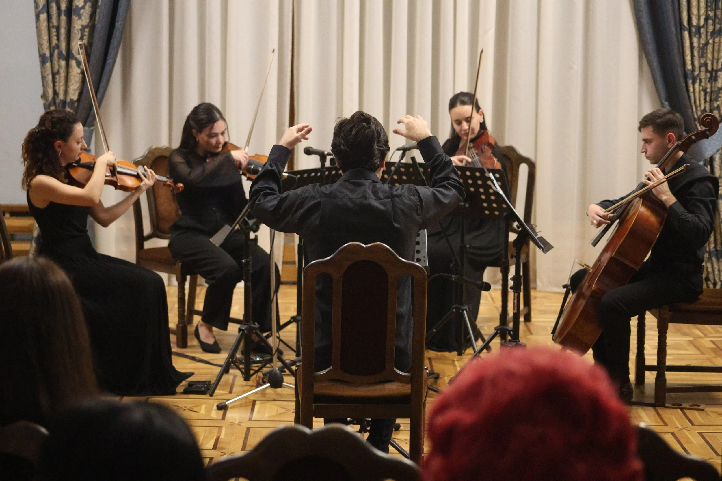 Orchestra performance featuring four string players and a conductor on stage in a concert hall with curtains.