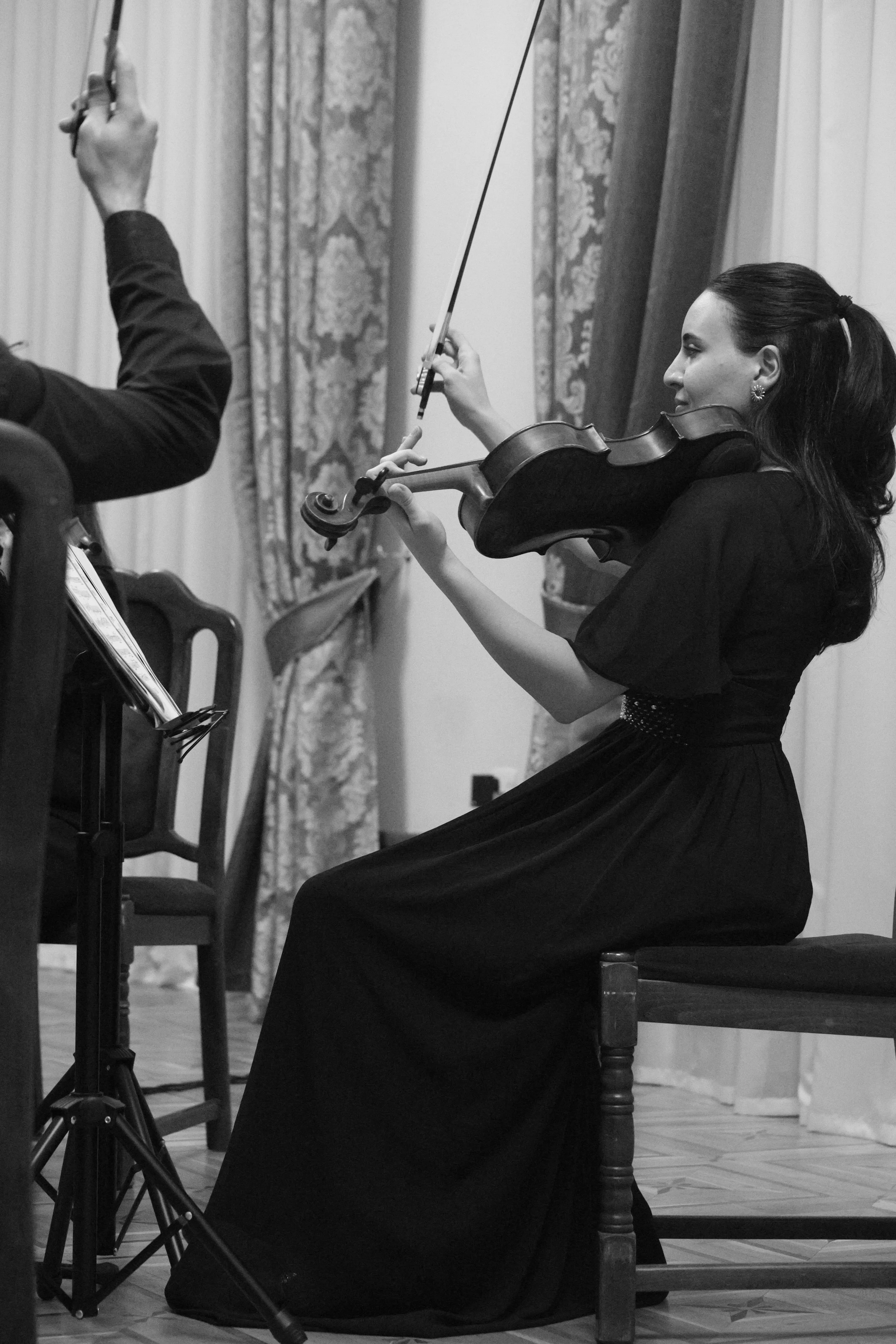 A woman playing the violin in a formal setting, sitting on a chair, with curtains and patterned floor tiles in the background.