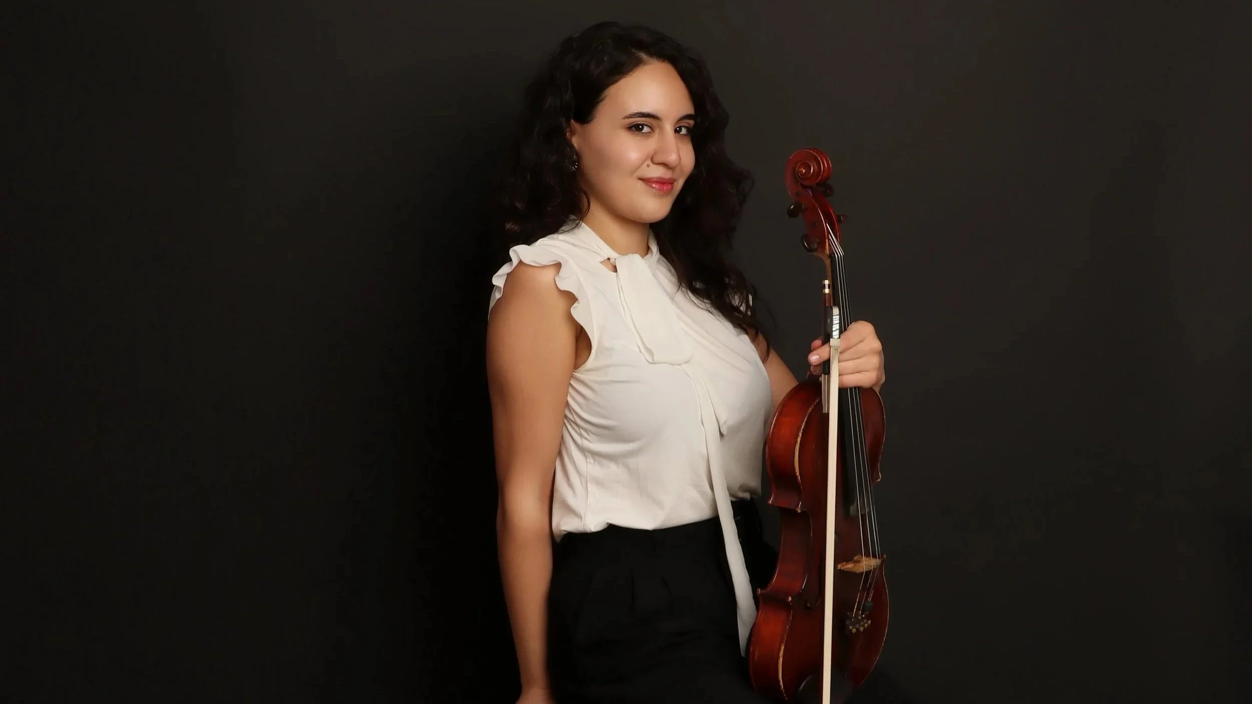 A woman with dark, curly hair and a confident expression, holding a violin and standing against a black background, wearing a sleeveless white blouse with ruffled shoulders.