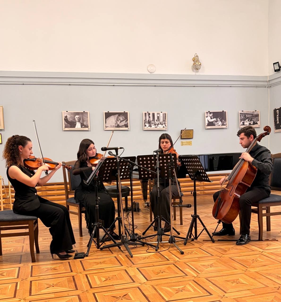 A string quartet performing in a room with wood flooring. The group includes two women playing violins, another woman playing viola, and a man playing cello. They are seated in front of music stands with sheet music, and there are black and white photographs hanging on the wall behind them.