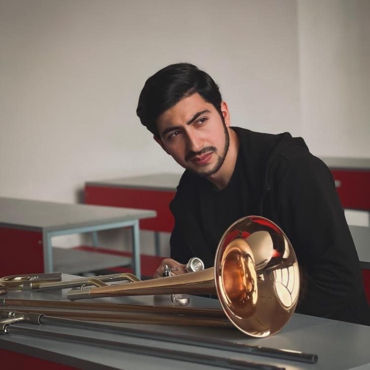 A young man with dark hair and a beard sitting at a classroom desk with musical instruments, including a tuba and trombone, on the desk.