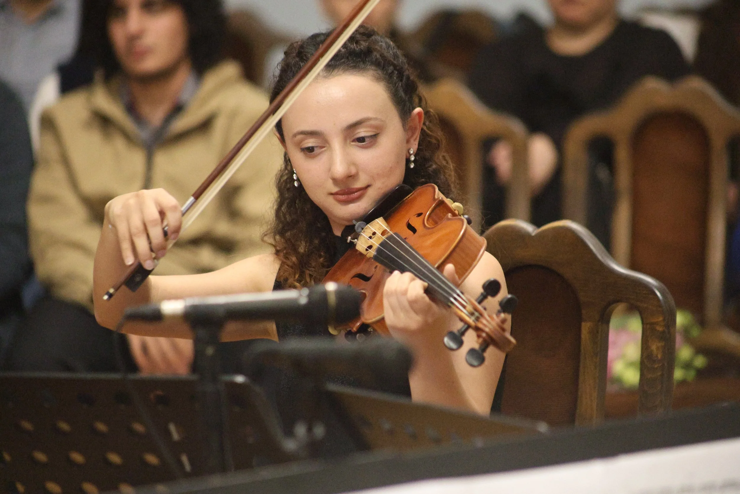 A young woman playing the violin during a performance, with an audience seated on wooden chairs in the background.