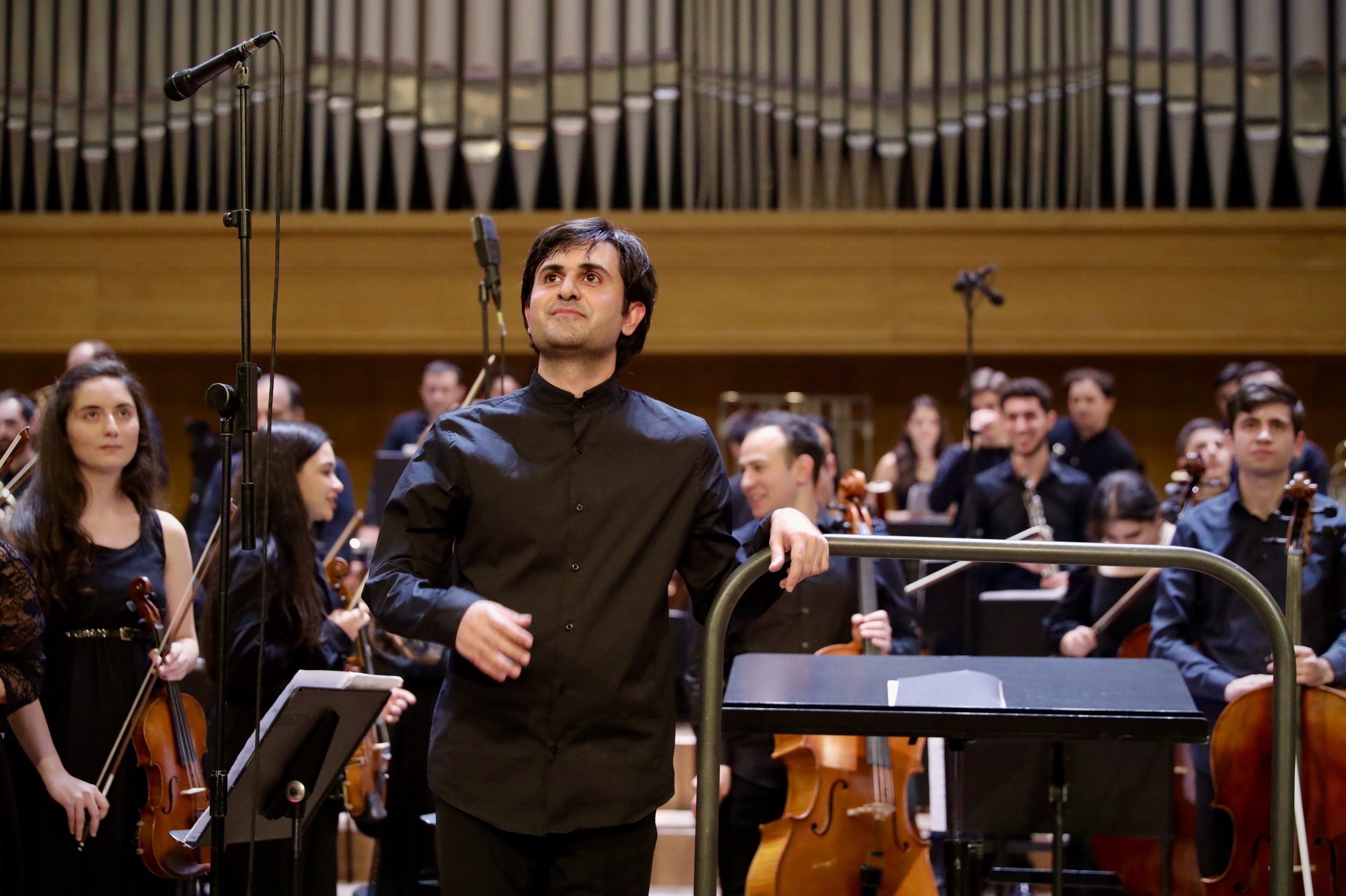A man in a black shirt stands in front of an orchestra holding a baton or conducting stick, with other musicians playing string instruments behind him in a concert hall.
