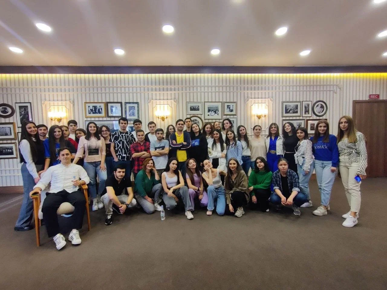Group photo of young people and an older man in a room with framed pictures on the wall and warm lighting.