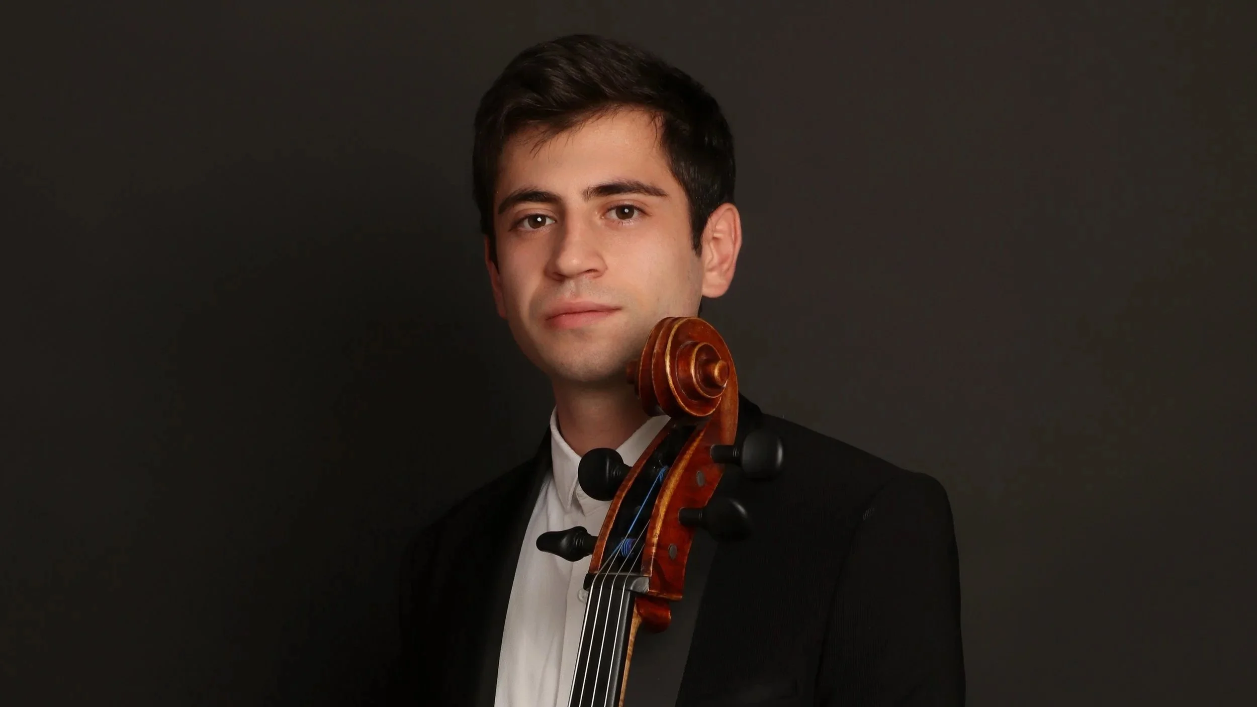 A young man with dark hair and light skin holding a cello, wearing a black suit and white shirt against a dark background.