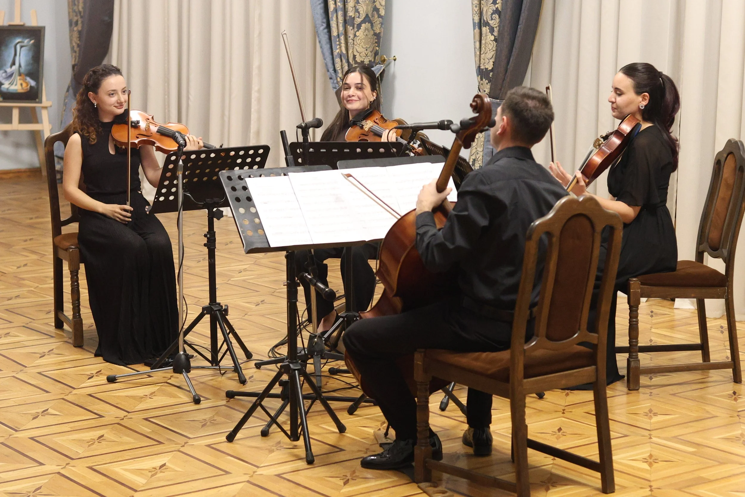 Four young musicians playing string instruments, including violins and a cello, during a performance in a room with ornate curtains and wood flooring.