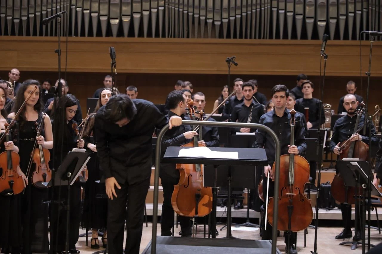 Orchestra members, including violinists, cellists, and wind players, on stage after a performance in a concert hall.