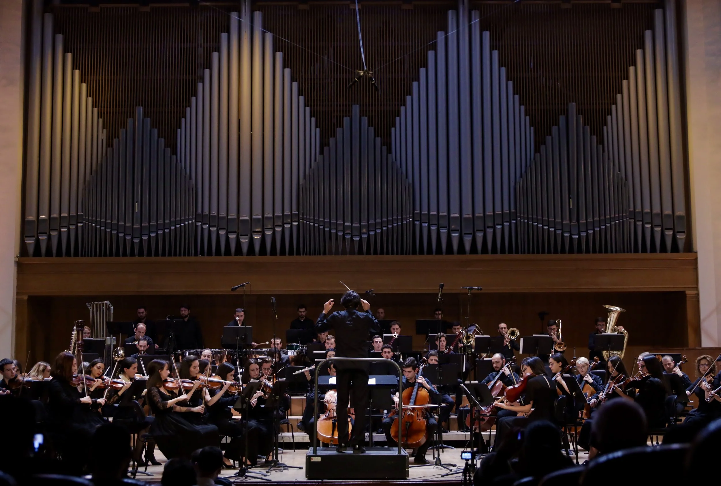 Orchestra performing on stage with wooden backdrop and large pipe organ in the background, conductor leading the musicians, audience seated in front.