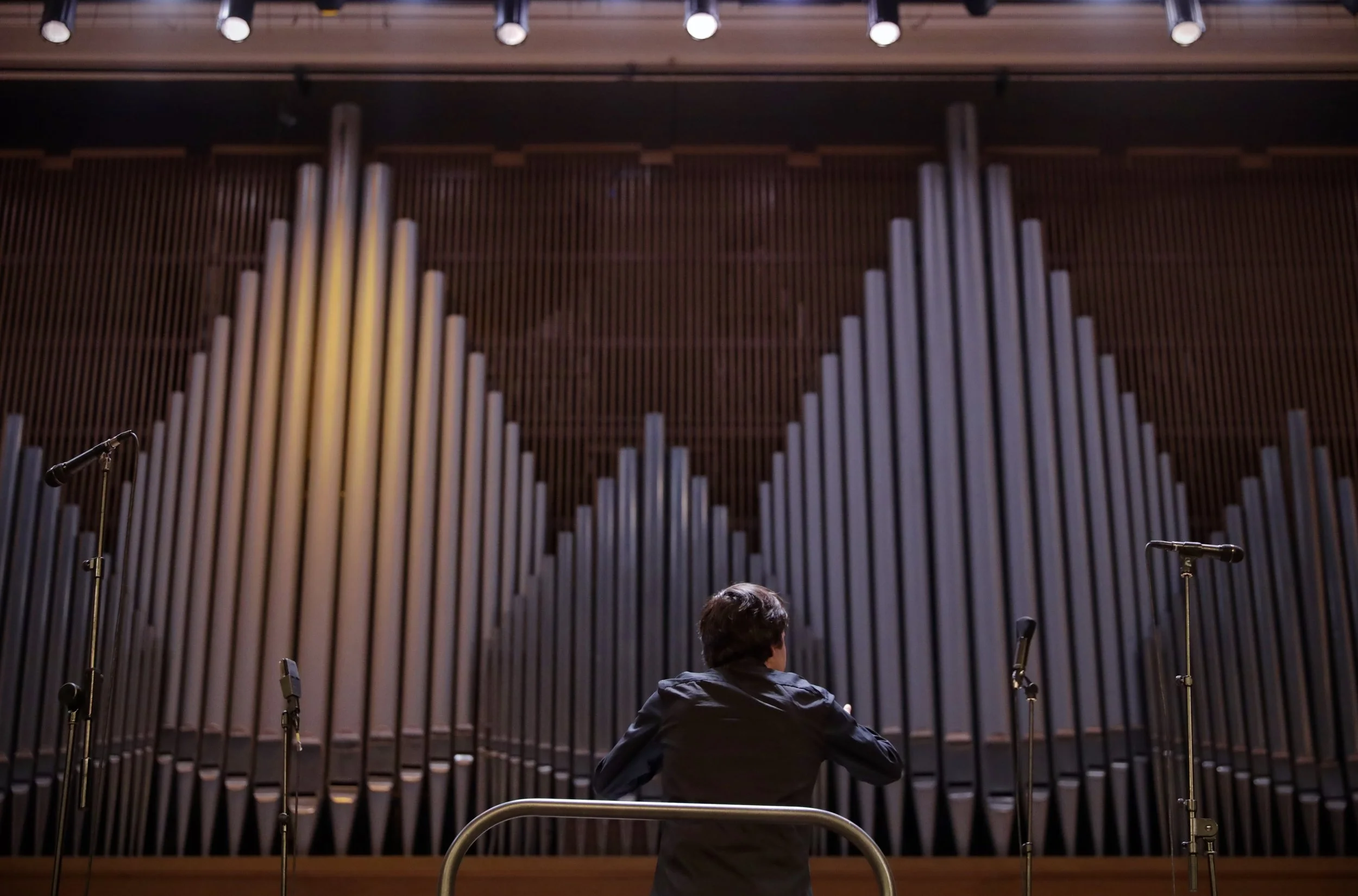 A man with dark hair sitting facing away in front of large organ pipes, with a wooden panel background and several microphones on stands nearby.