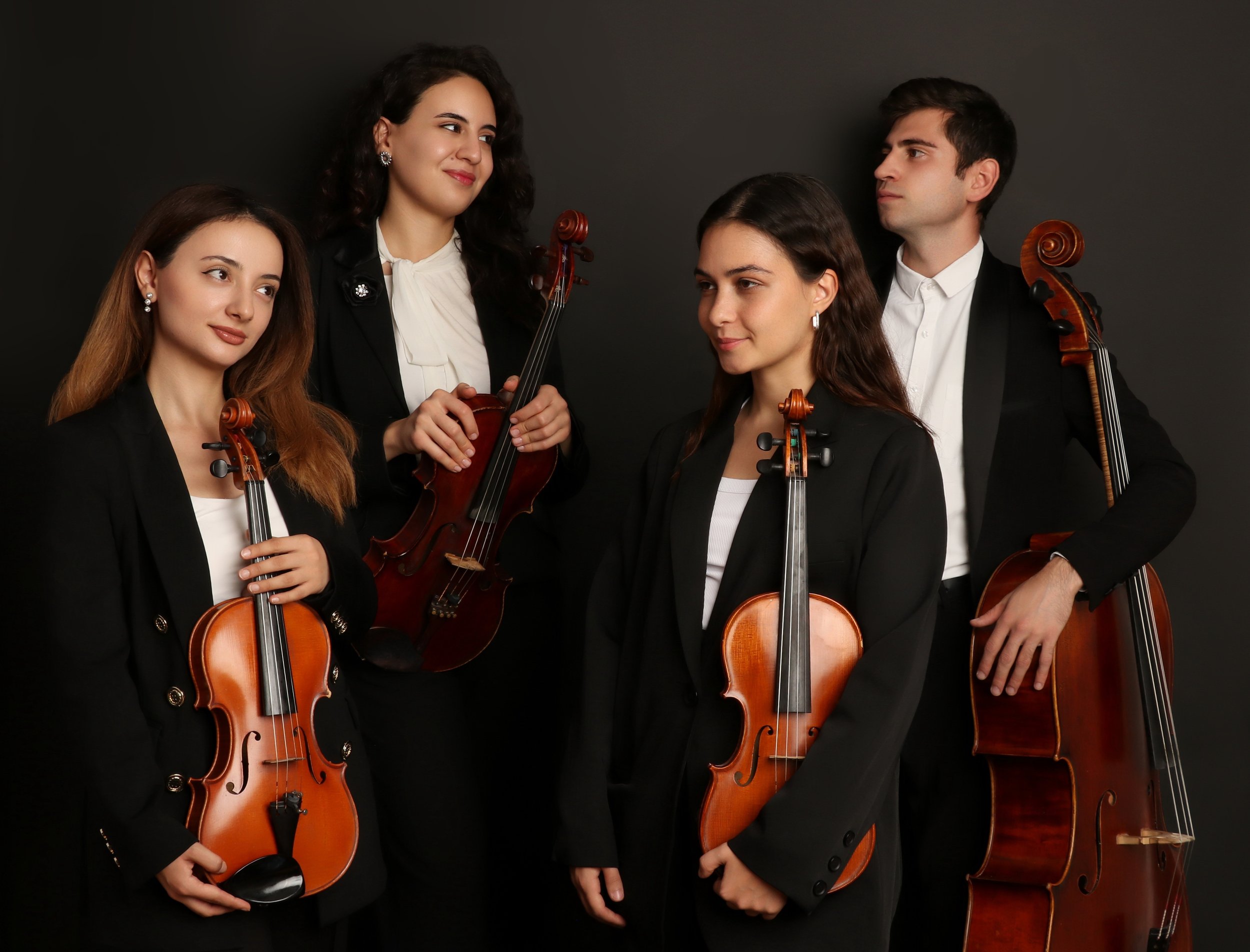 A group of five young adults dressed in black suits holding violins and violas, standing against a dark background.