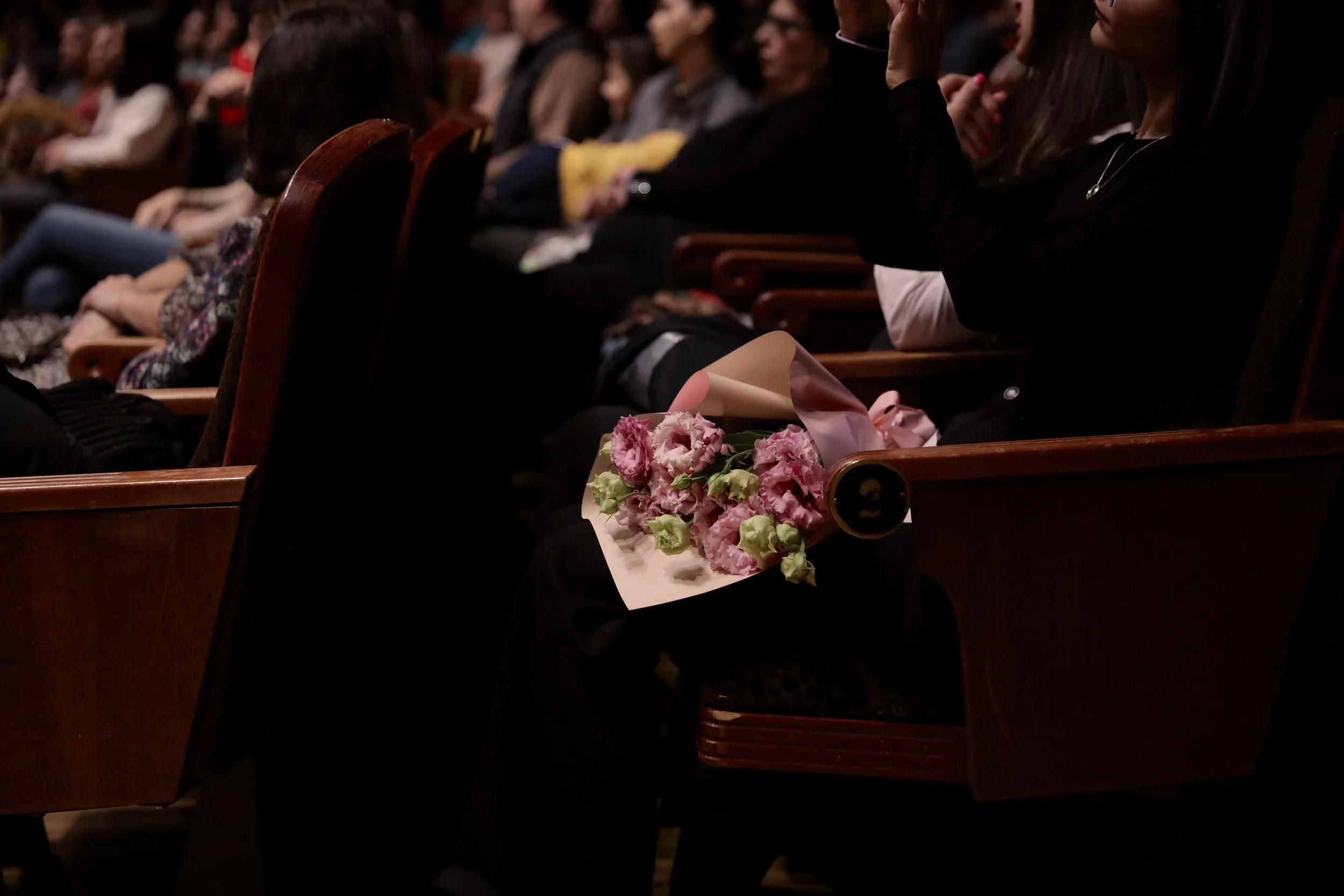 Audience seated in theater with a woman holding a bouquet of pink flowers.