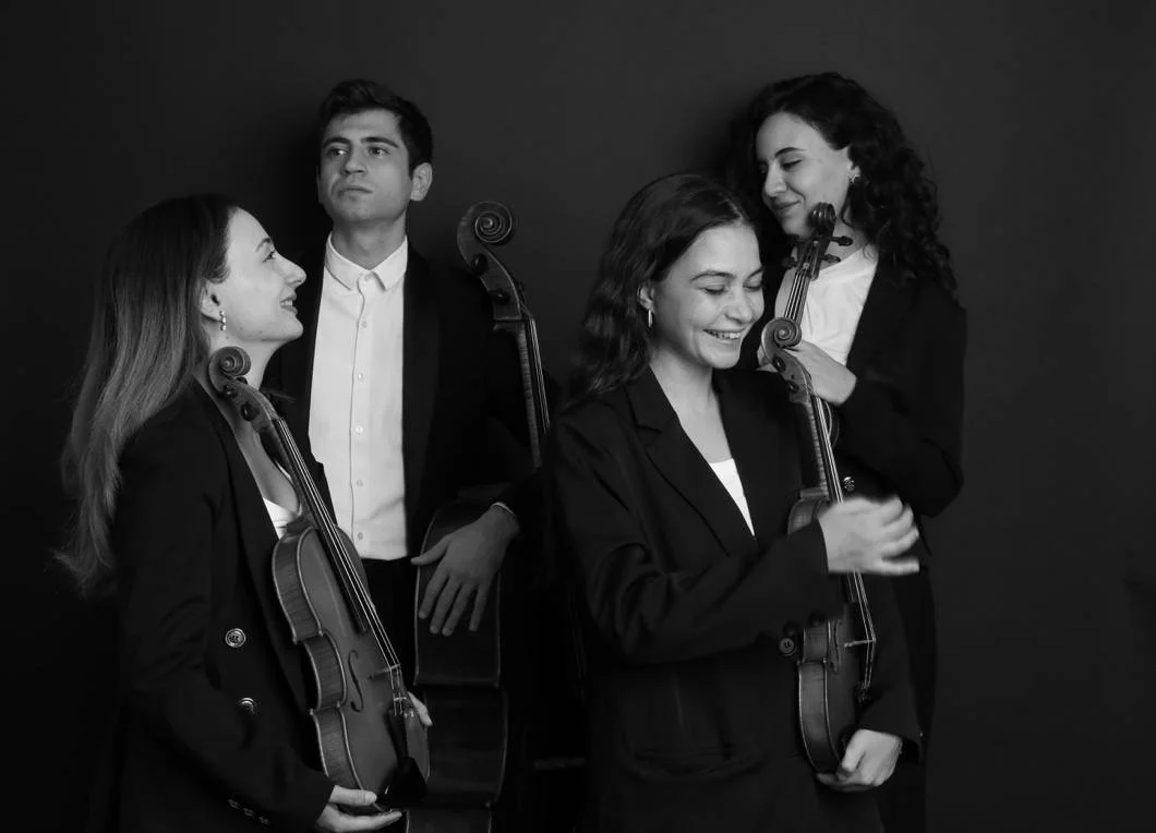 Four musicians holding violins, dressed in formal black attire, standing against a dark background.