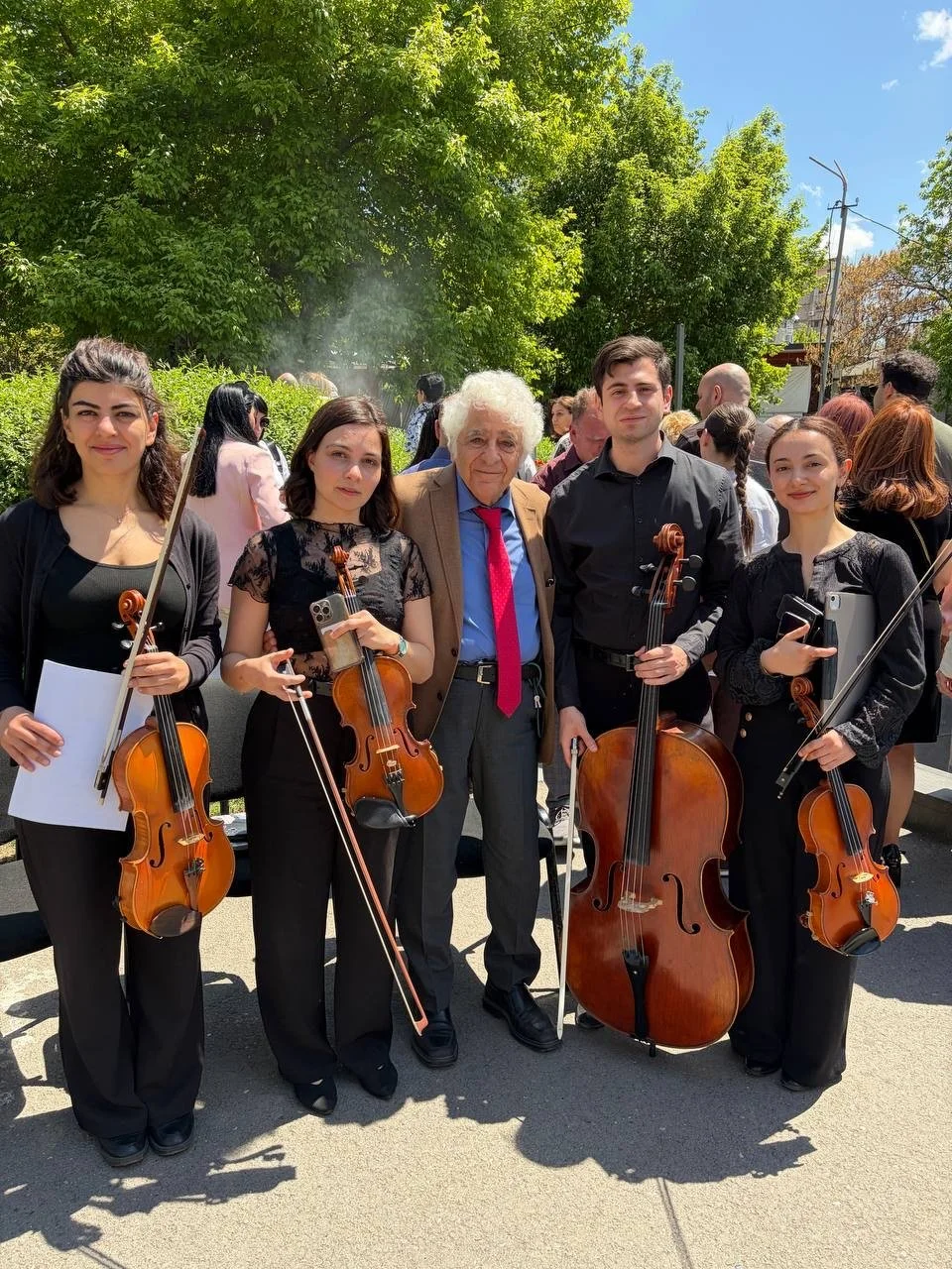 Group of five musicians, four women and one elderly man, holding string instruments like violins and a cello, standing outdoors with a crowd and green trees in the background.