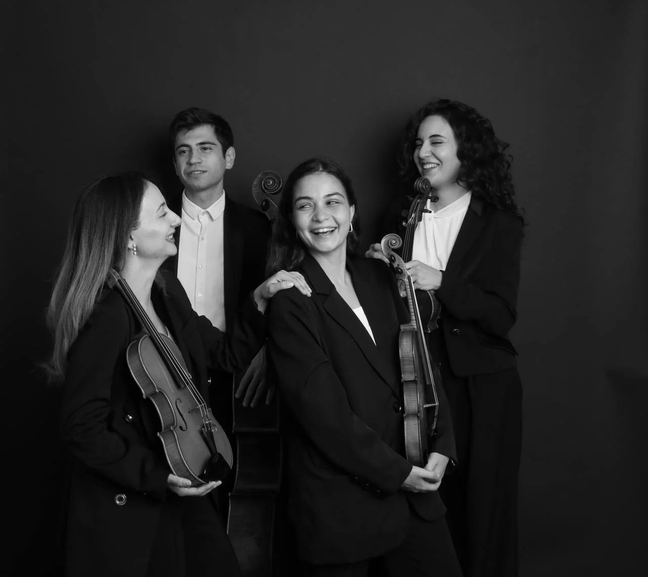 Four musicians, three women and one man, stand together holding violins, and are dressed in formal black attire. They are smiling and laughing, engaging with each other, against a plain dark background.