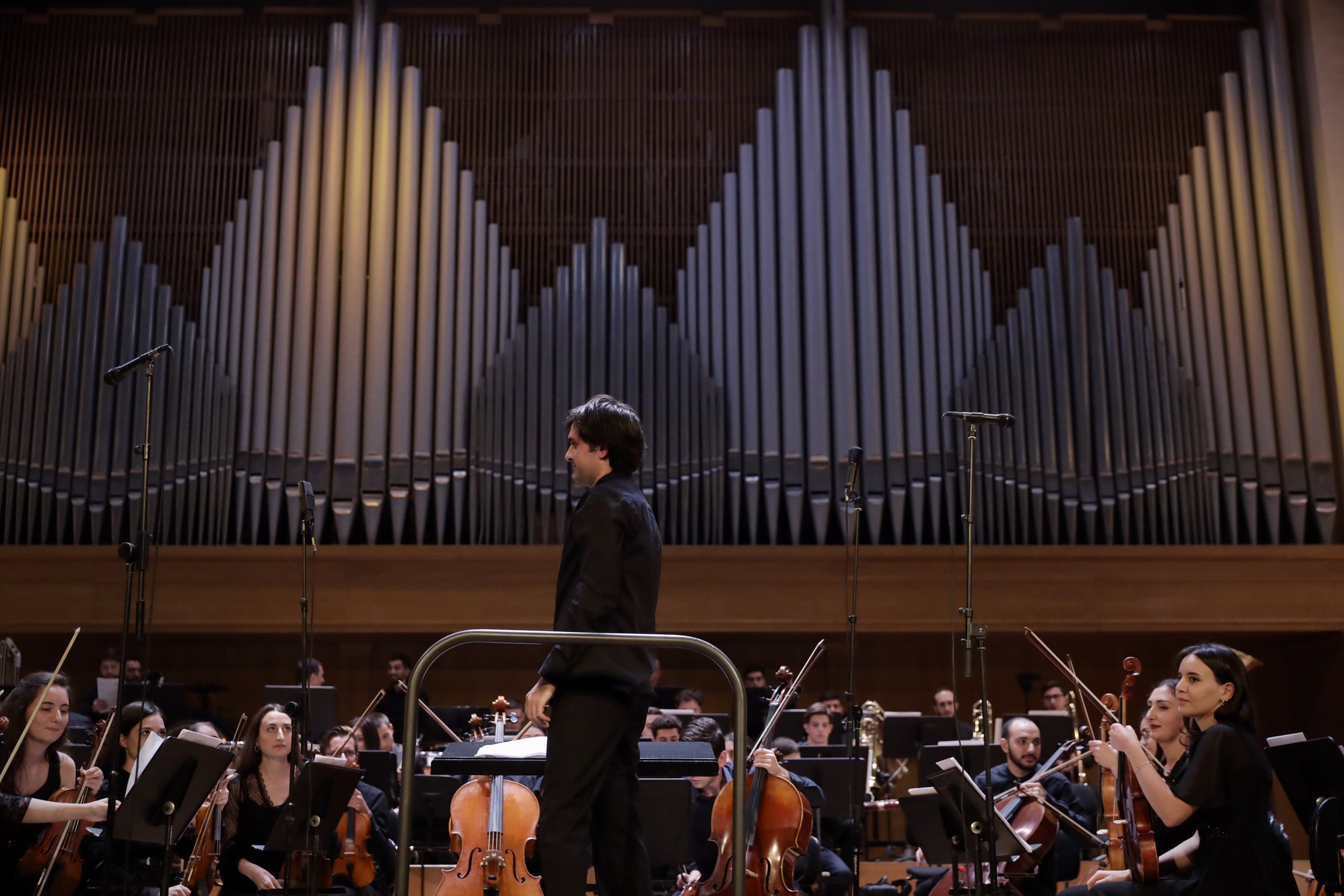 Man in black tuxedo leading an orchestra in a concert hall with a large organ in the background.