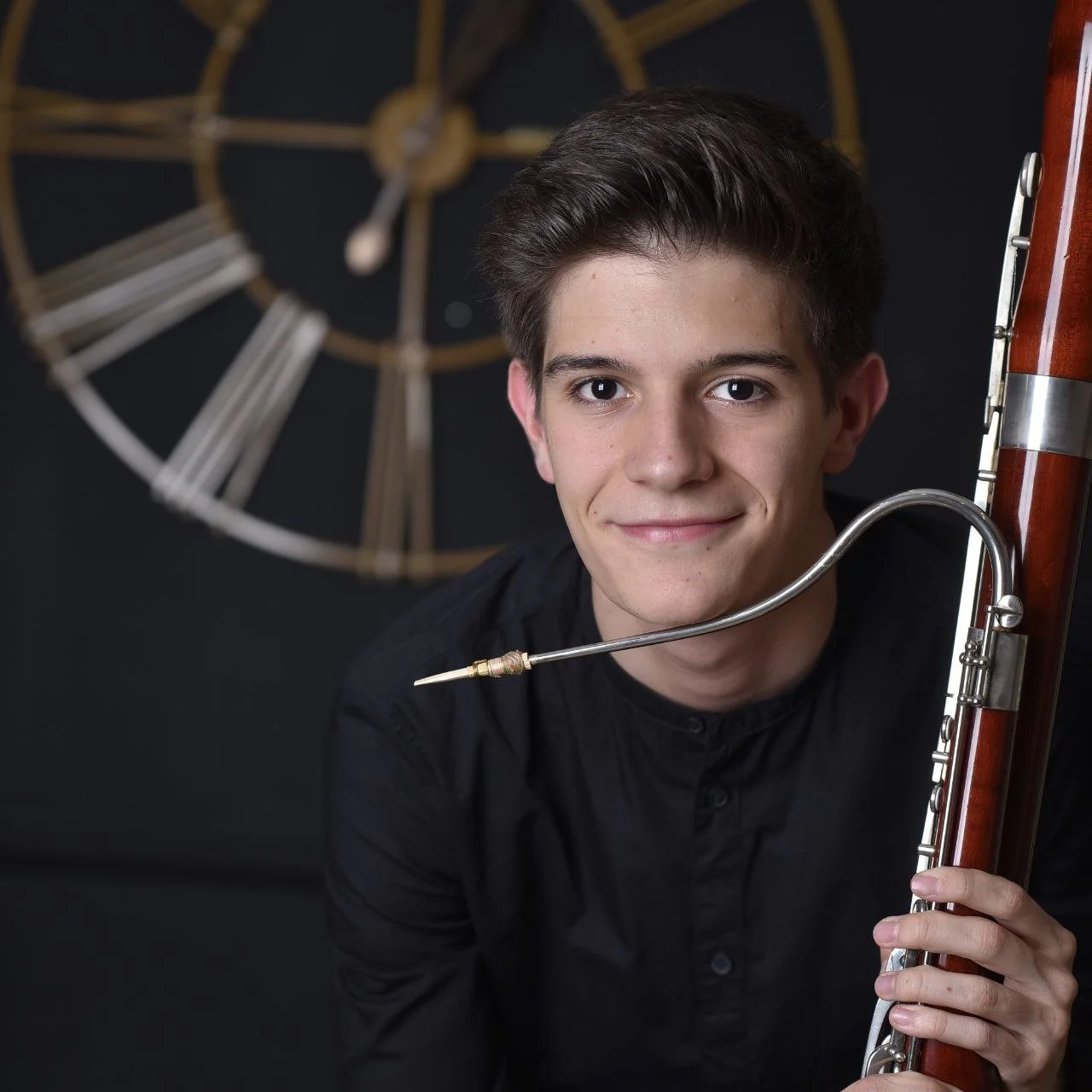 A young man holding a bassoon, with a large clock in the background.