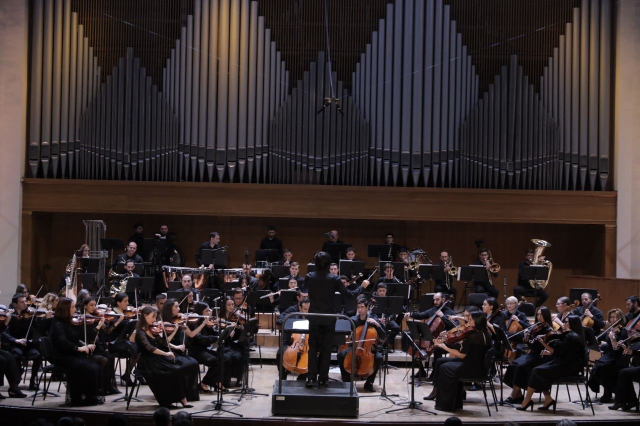 An orchestra performing on stage in a concert hall with a conductor in front, large pipe organ in the background.