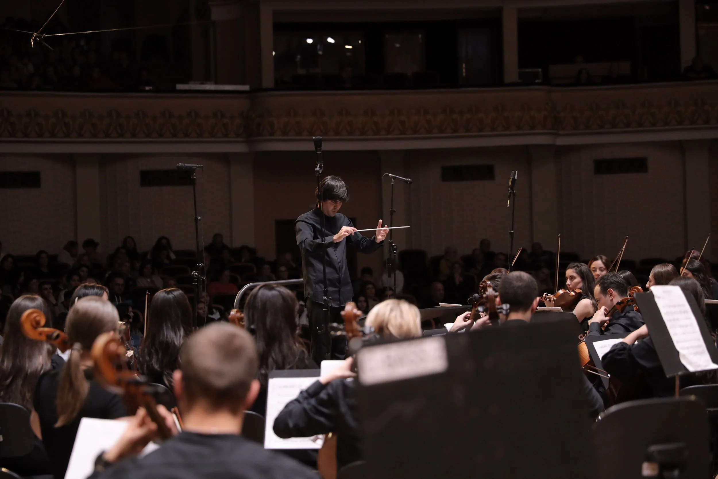 A conductor leading an orchestra on stage during a performance, audience watching in the background.