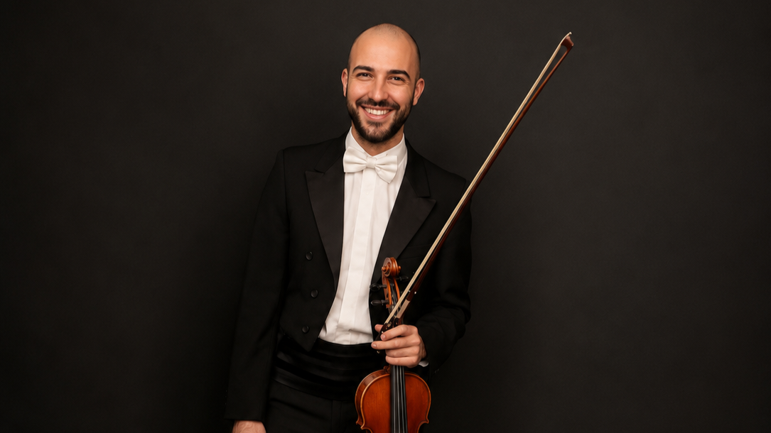 A man dressed in a tuxedo and bow tie holding a violin and smiling against a dark background.