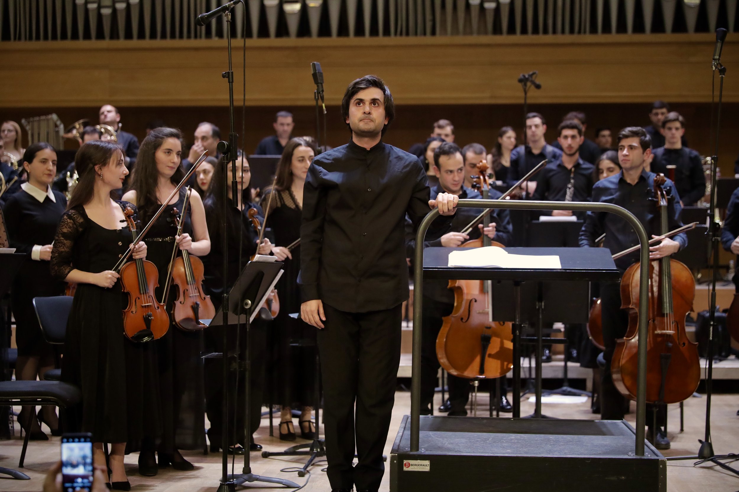 Orchestra conductor in black standing in front of an orchestra in a concert hall.