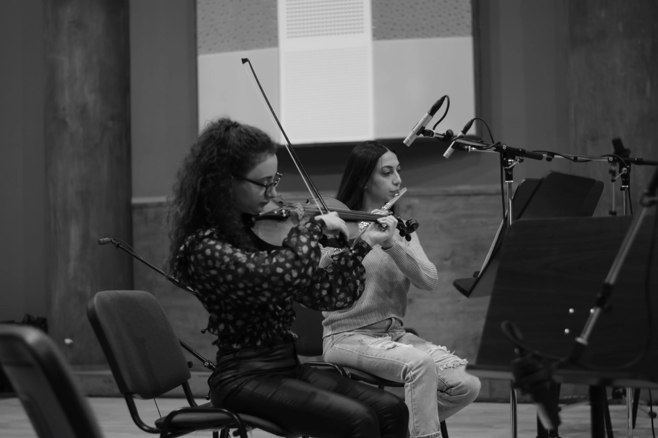 Two women are playing violins while sitting on chairs in a recording studio, with microphones positioned in front of them and a large screen displaying sound waves in the background.