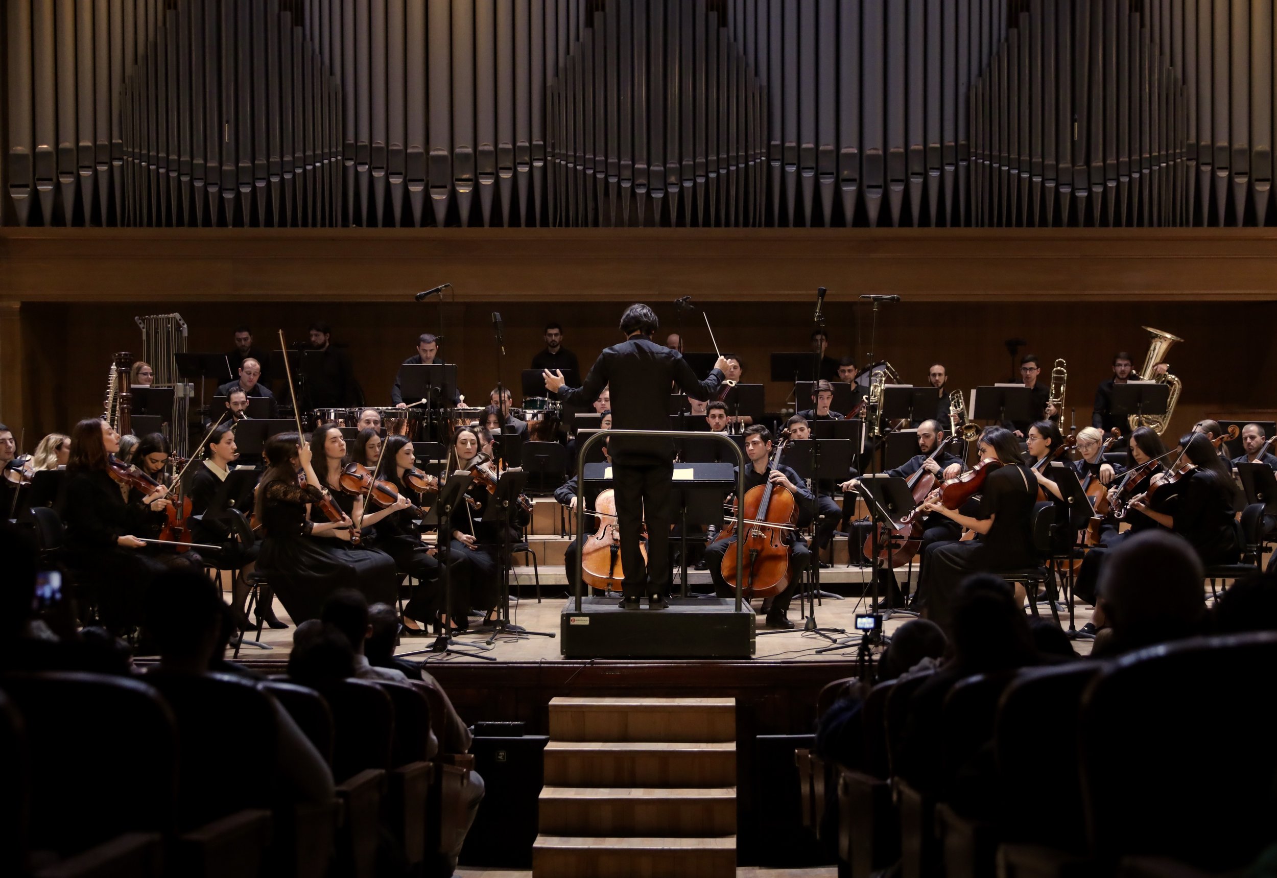 Orchestra performing on stage with conductor leading and audience in front.