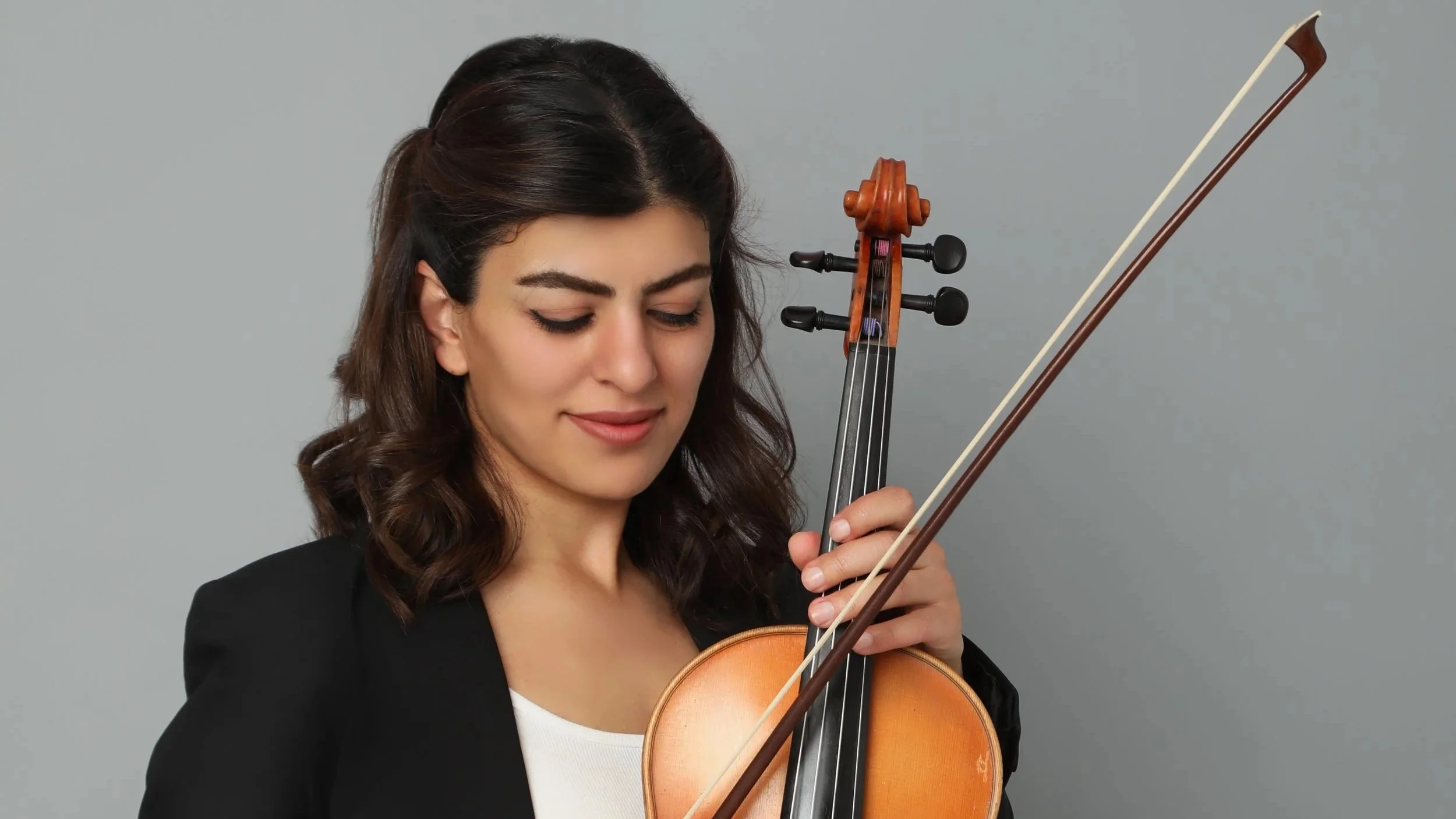 Woman with dark hair playing a violin against a plain gray background.